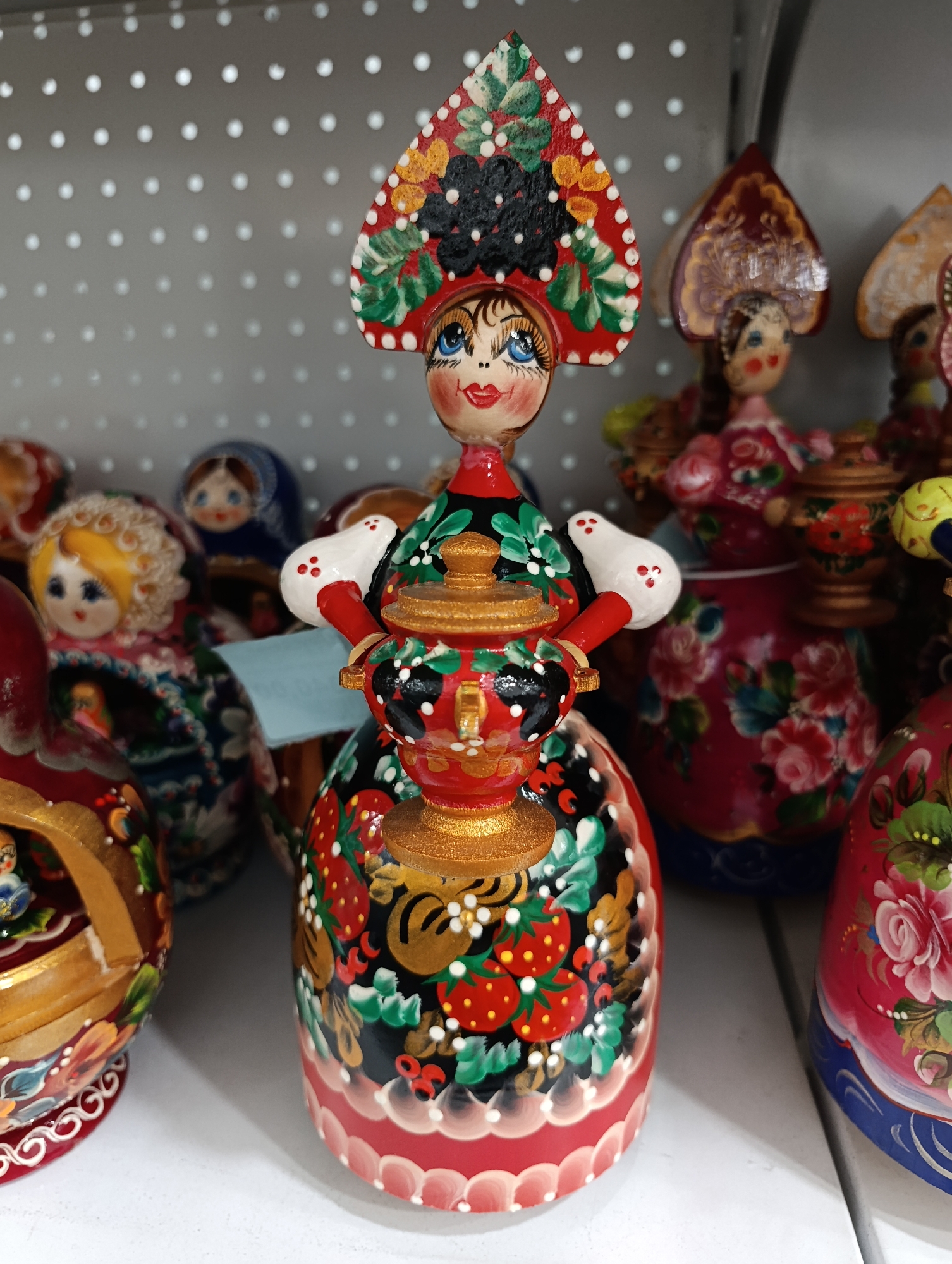 The image captures a vibrant display of traditional Russian nesting dolls (matryoshka), likely in a souvenir shop in Birobidzhan, Russia.
The central subject is a tall, exquisitely detailed matryoshka doll, painted in rich folk art style. It features a woman with bright blue eyes, rosy cheeks, and red lips, wearing a red kokoshnik-style headpiece adorned with black and yellow berries, green leaves, and white dots. Her dress is predominantly black with red and green sections, intricately decorated with strawberries, green foliage, and tiny white flowers. Uniquely, this doll holds a small, golden samovar, a traditional Russian tea urn, in its hands. The base of the doll is a scalloped red pattern with white accents.
In the background, an array of other matryoshka dolls is visible, showcasing similar craftsmanship and varying designs. These dolls feature floral patterns, such as pink roses and green leaves, on bodies of red, pink, and blue, with some sporting different kokoshnik styles. One doll in the mid-left background has a light blue tag attached, but the text is unreadable. The dolls are arranged on white, perforated shelving, characteristic of a retail display. The scene is brightly lit, emphasizing the intricate hand-painted details and vibrant colors of the traditional Russian folk art. No specific activity is occurring; it is a static display of cultural artifacts.