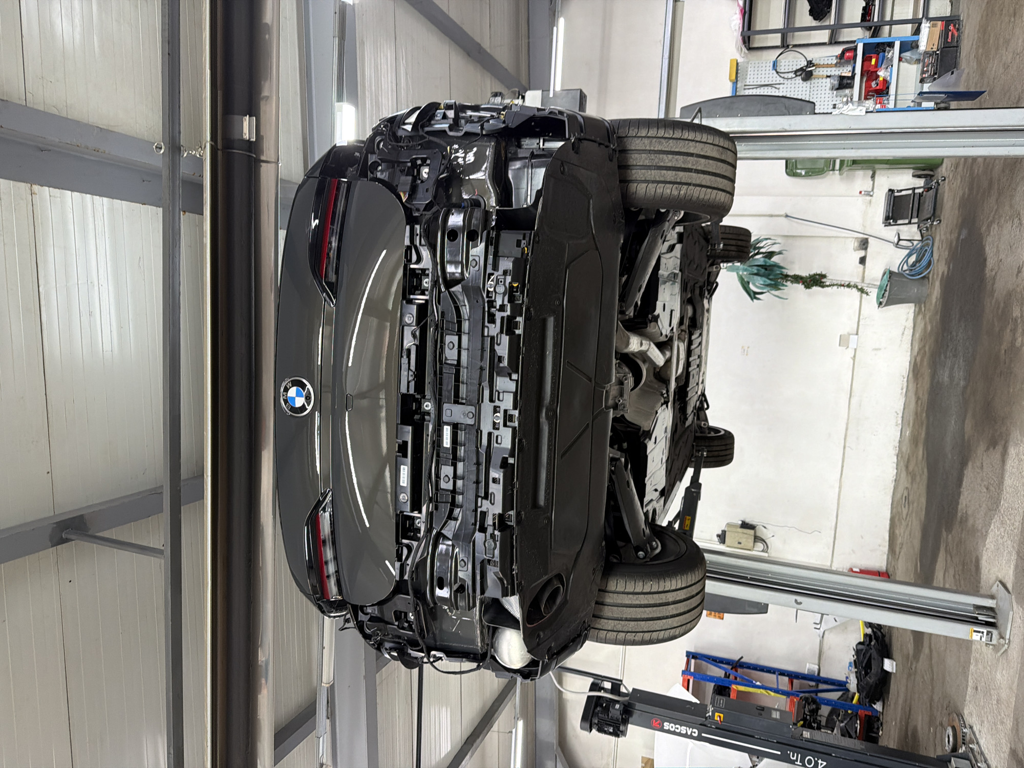 A dark gray BMW car is prominently featured, elevated on a hydraulic car lift within what appears to be a professional service garage in Burgas, Bulgaria. The car is seen from its underside, revealing the chassis, suspension components, wheels with tires, and parts of the exhaust system, including a muffler and pipes. The BMW logo is clearly visible on the rear trunk.
The setting is a well-lit indoor space, likely a workshop. The walls are made of light-colored corrugated metal panels, and the floor is concrete. In the background, various garage equipment and tools are visible, including a blue and red tool chest, a pegboard with items hanging on it, and what looks like a tire alignment sensor. A blue hose reel and other miscellaneous items are arranged on the floor. Unusually for a garage, a green decorative plant in a pot is also present.
The car lift itself is a prominent feature, with the brand "CASCOS" and a capacity of "4.0 Tn." clearly printed on its arm. The car's underbody appears relatively clean and intact, suggesting routine maintenance or inspection rather than significant damage. There are no people visible in the image, but the setup indicates an ongoing or recent automotive service activity.