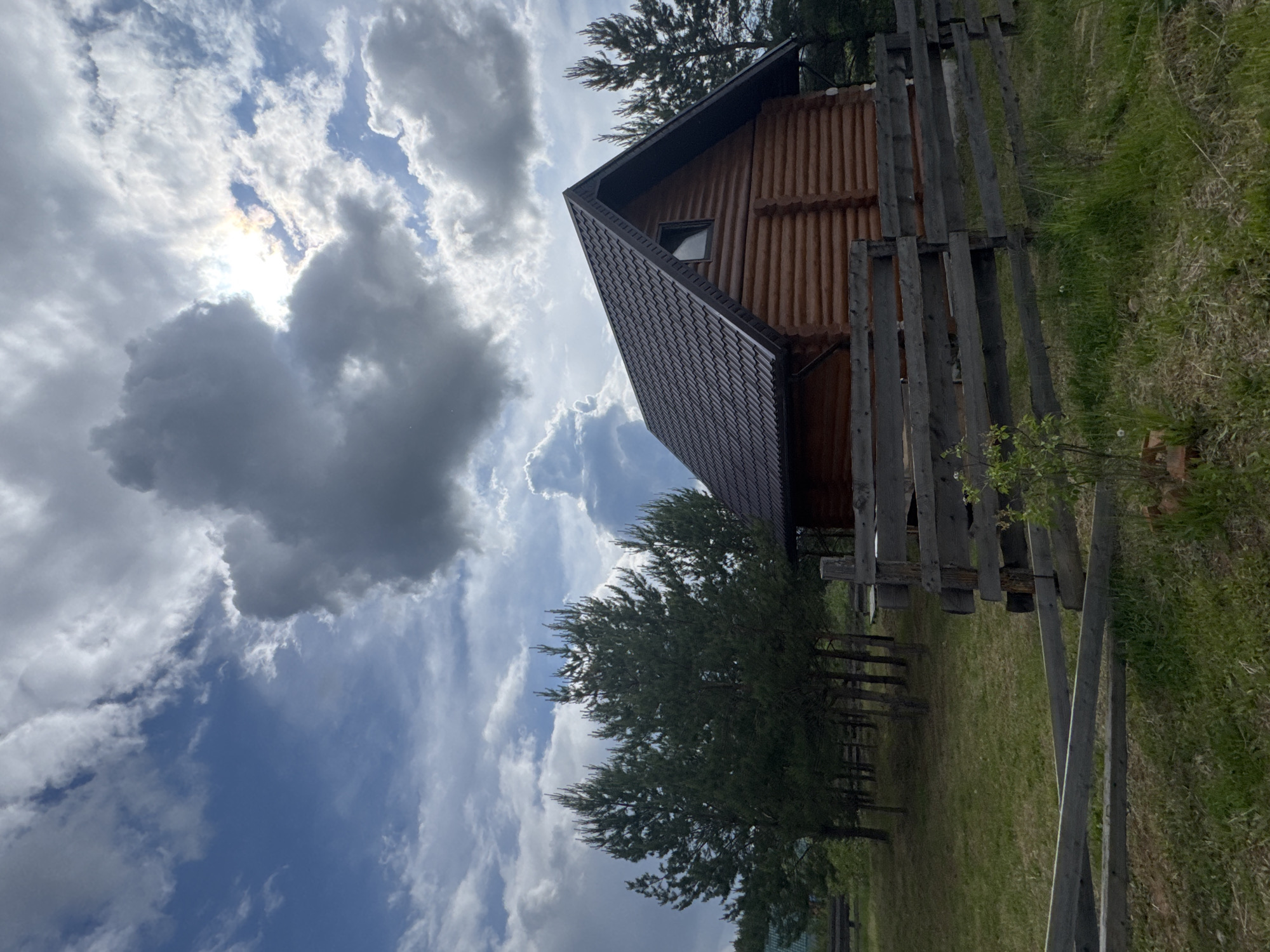 The image depicts a wooden cabin with a dark, gabled roof, situated on a grassy slope and enclosed by a rustic wooden fence. The sky, which occupies a significant portion of the frame, is filled with dynamic cloud formations, with the sun partially obscured. Some evergreen trees are also present. No people are visible. The location cannot be determined definitively without further information.