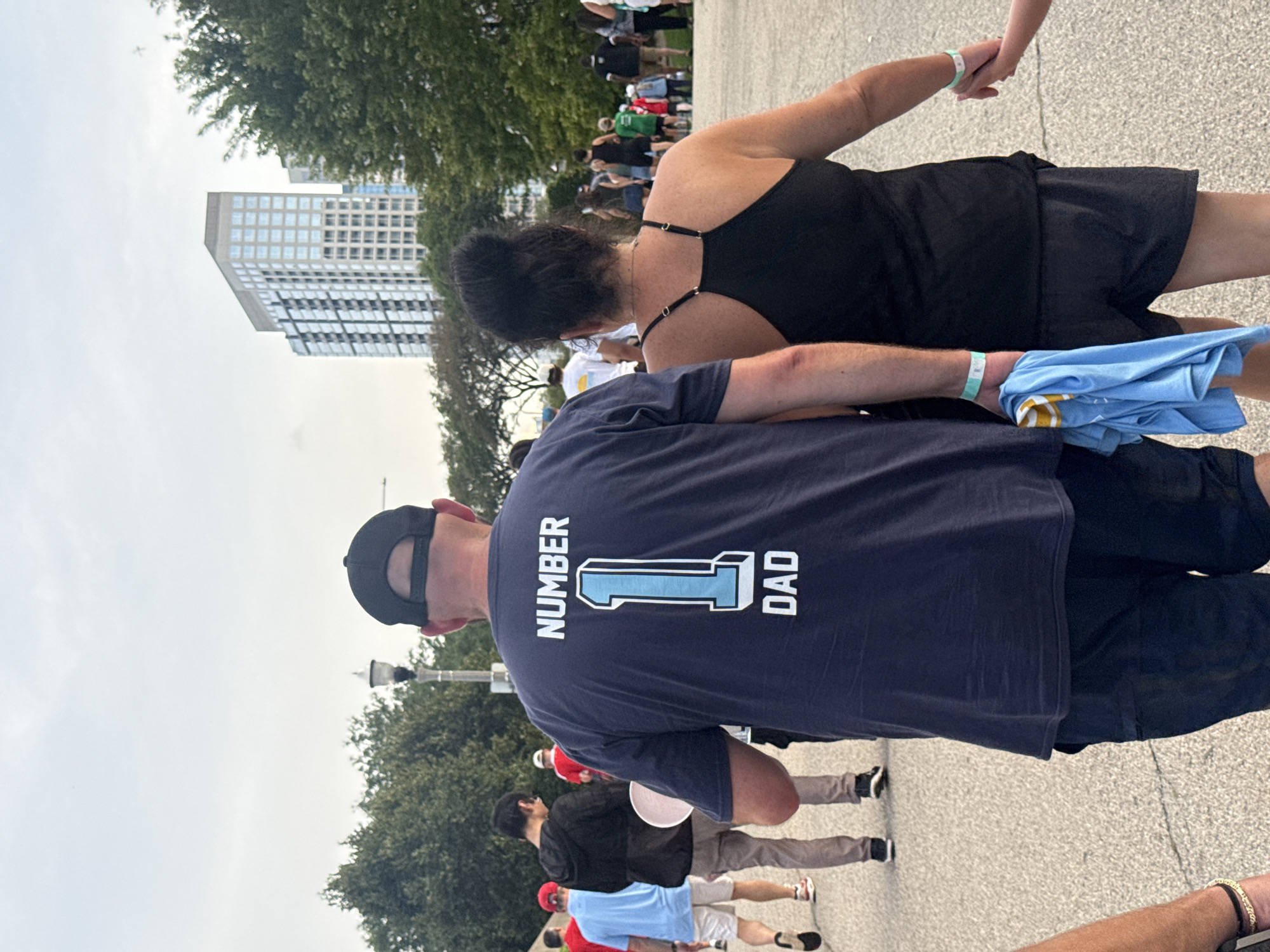 The image shows a rear view of a man wearing a dark blue t-shirt with "NUMBER 1 DAD" printed in white and light blue on the back. He is wearing a black baseball cap and dark pants. He is holding a light blue t-shirt in his left hand. To his right, a woman in a black tank top and black shorts is holding hands with a child, whose arm is visible at the edge of the frame. They are walking on a paved path, possibly in a park. In the background, there are trees, a tall modern building with many windows, and other people walking in the distance. The sky is overcast. The overall scene suggests a public event or gathering in an urban park setting.