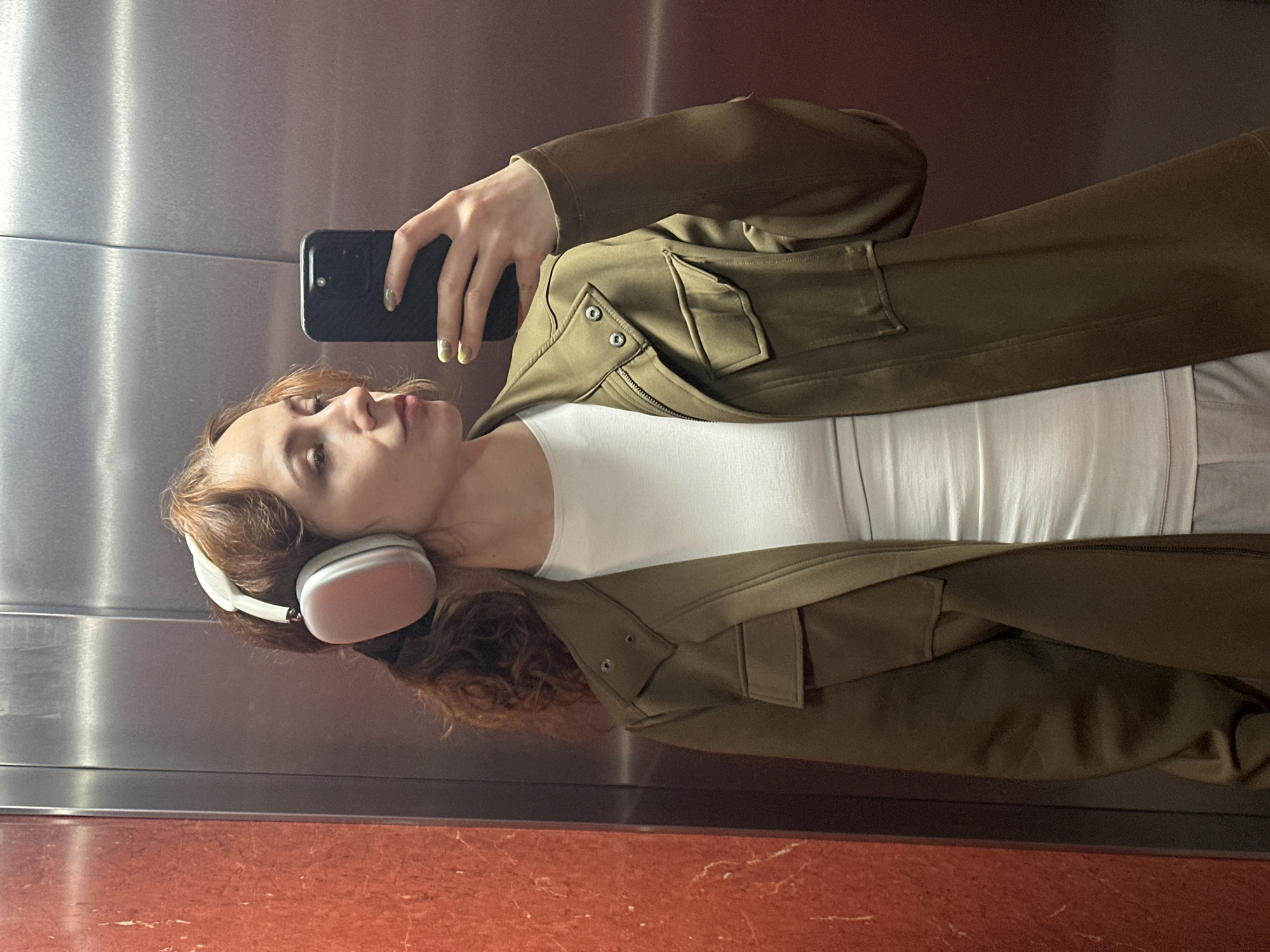 A woman takes a selfie in an elevator. She wears white headphones, a white t-shirt, and a green jacket. She holds a black smartphone in her right hand. The elevator has a brushed metal wall and a red marble floor. The location is Novi Sad, Serbia, based on the red marble floors in common elevators there.