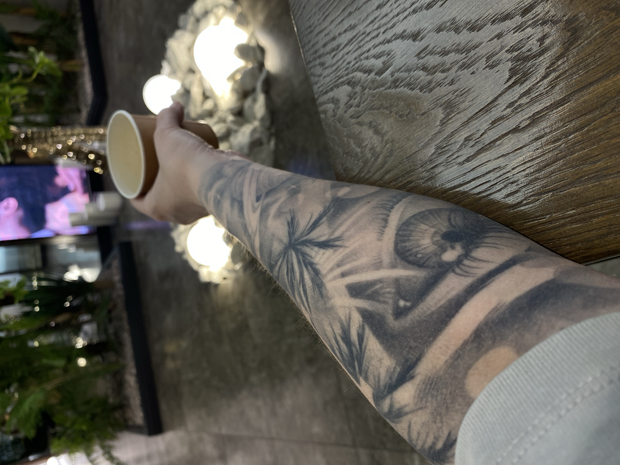 A close-up captures an arm with a grayscale tattoo, adorned with a stylized eye and botanical patterns, holding a brown paper cup. In the background, a television displays an indistinct image of a person, framed by a dark structure with plant life. Further back, blurred lights are visible. A dark wooden surface is present at the top right. The photo's location is not specified, and therefore cannot be stated with confidence.