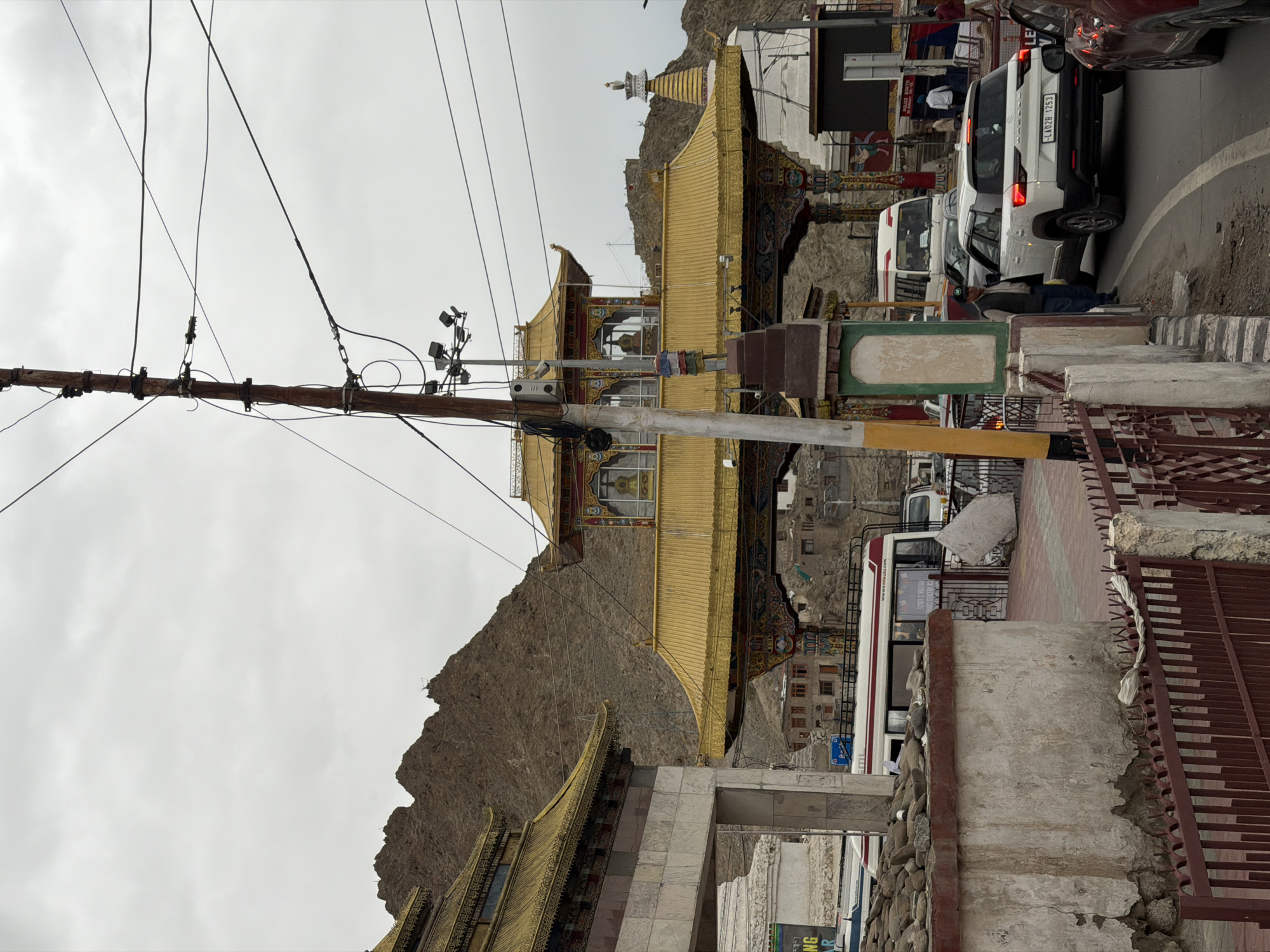 The image depicts a street scene in Leh, India, with a prominent ornate gateway featuring golden roofs and intricate paintings. The gateway appears to be a significant cultural or religious structure, possibly leading to a monastery or temple complex, set against a backdrop of rugged, brown mountains under a cloudy sky.
In the foreground, a white SUV with the license plate "LA02B 1253" is stopped on the right side of the road. A man in a cap and vest stands near the open driver's side door of the SUV, facing towards the road. To the left of the SUV, a section of a red metal fence is visible. Beyond the SUV, a red car is partially visible.
Further into the scene, a white bus with red and blue stripes is parked or moving slowly in traffic on the left side of the road. The bus has advertisements on its side. A power pole with numerous wires stretches across the upper portion of the image, obscuring parts of the sky and the gateway.
The architecture of the gateway and the surrounding buildings suggests a blend of traditional and modern elements. The street appears to be a main thoroughfare with vehicles and pedestrians. The overall atmosphere is somewhat overcast, with muted lighting. The presence of traffic and infrastructure like the power pole indicates an urban or semi-urban environment. The mountainous landscape is characteristic of Leh's geographical setting.