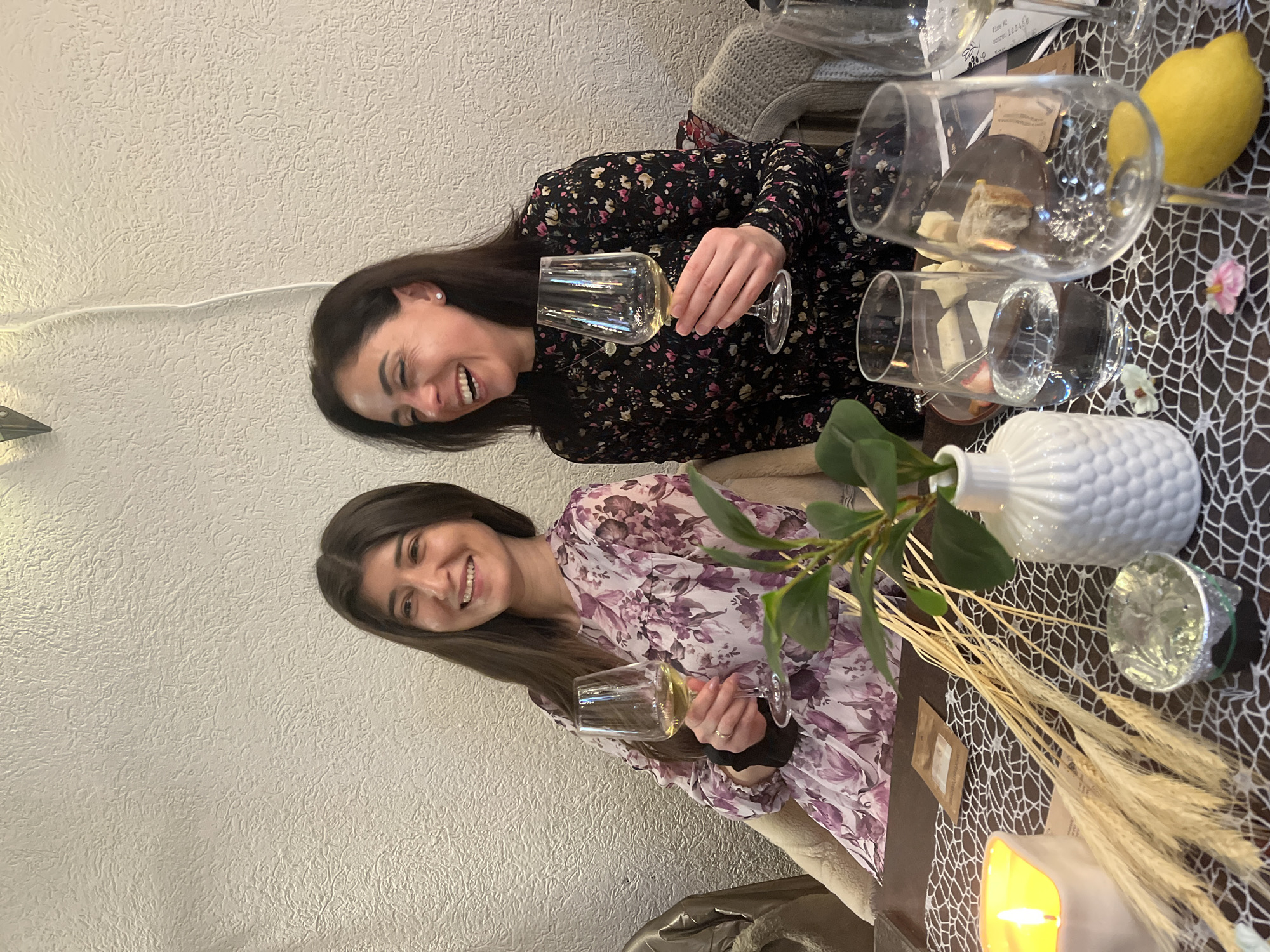 Two women are seated at a table in an indoor setting. The woman on the left wears a floral print dress and holds a wine glass. The woman on the right wears a darker floral print dress and is also holding a wine glass while smiling and laughing. The table has a decorative white net-like table runner. There are several wine glasses, a white vase with greenery, and wheat stalks used as decorations. A yellow lemon sits near one of the glasses. The environment appears to be a restaurant or event space. It is not possible to confirm that this picture was taken in Varna, Bulgaria based on the visible image alone.