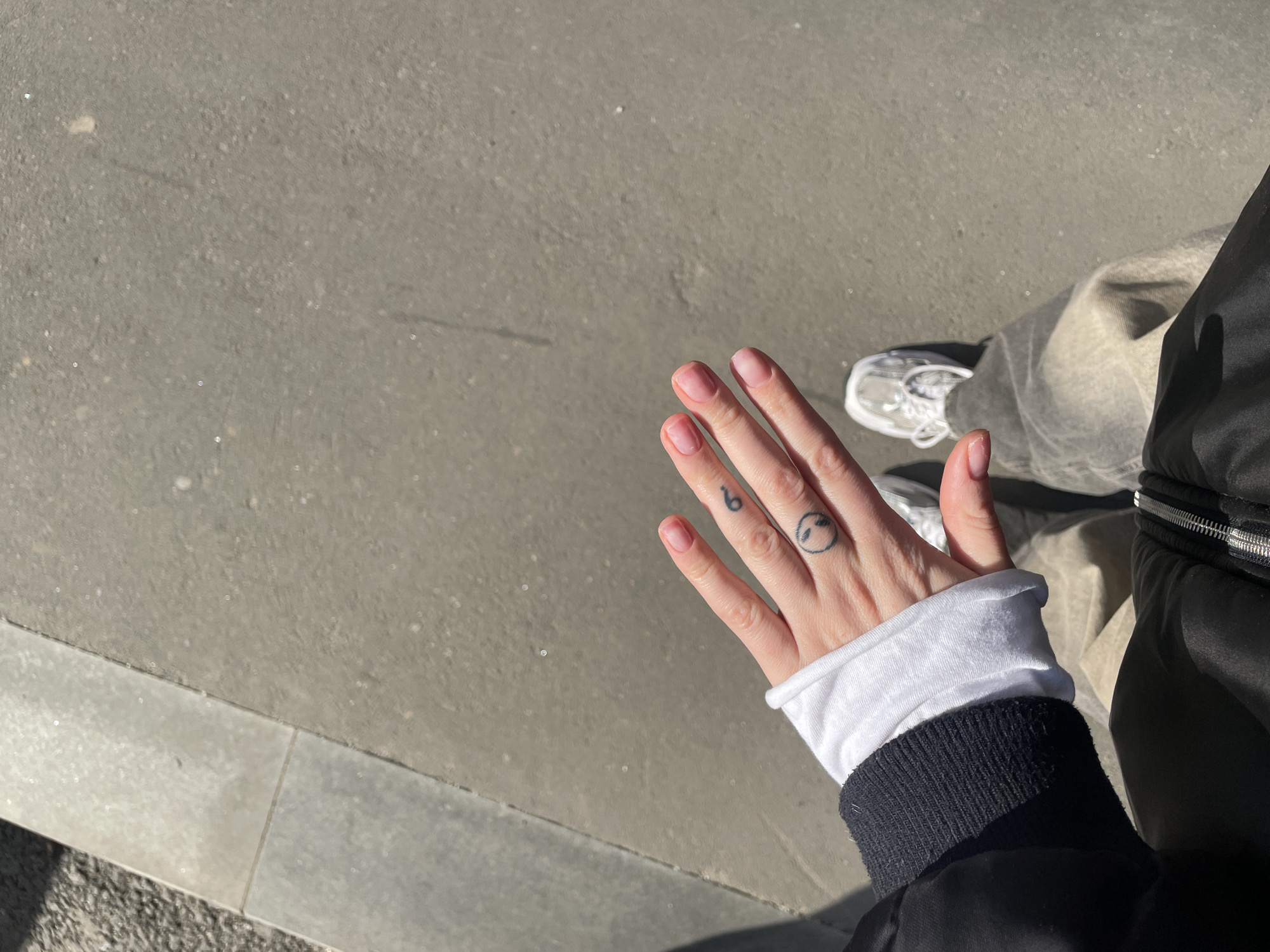 A first-person, downward-angle shot captures a person standing on a paved urban surface in Moscow, Russia, under bright daylight. The subject's right hand is prominent in the foreground, featuring short, light pink fingernails. Two small black tattoos are visible on the hand: the numeral '6' on the ring finger and a circular smiley face on the middle finger. The subject wears a black jacket with a visible zippered front and ribbed cuff, a white long-sleeved shirt layered underneath, light grey faded baggy pants, and light grey athletic sneakers with white laces. The ground consists of a lighter grey concrete or tiled border on the left, adjacent to a darker, textured asphalt or paved area that occupies the majority of the frame. A shadow, likely from the person's upper body, is cast onto the ground in the lower-left corner.