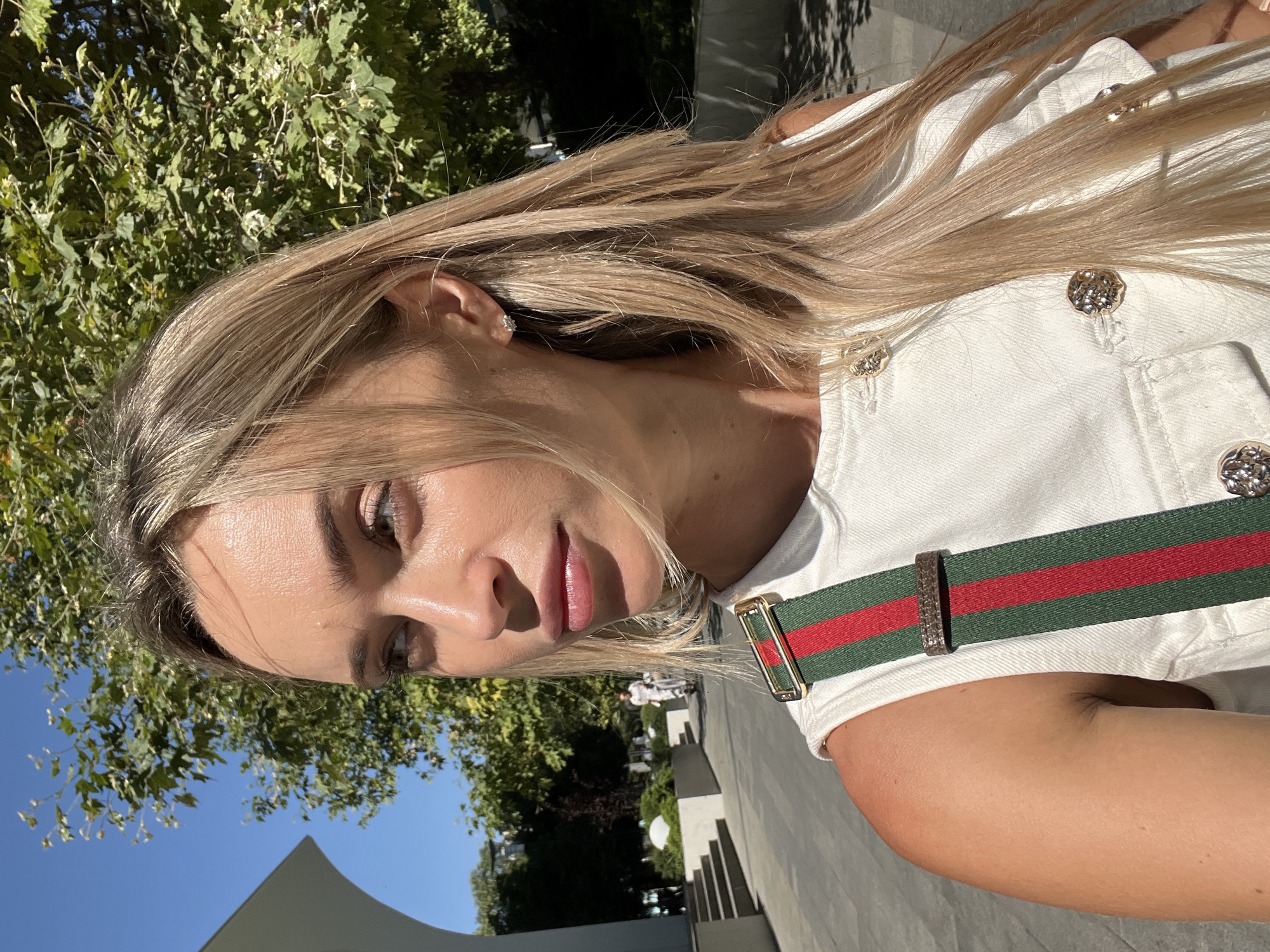 A woman with blonde hair is pictured from the chest up, looking to the left. She is wearing a white top with gold buttons and a green and red striped strap across her shoulder. She has a diamond earring in her left ear. The background is a sunlit outdoor area with trees, a blue sky, and a building with an arched entrance. In the distance, a person on a bicycle is visible. The location is the city of Odesa, Ukraine.