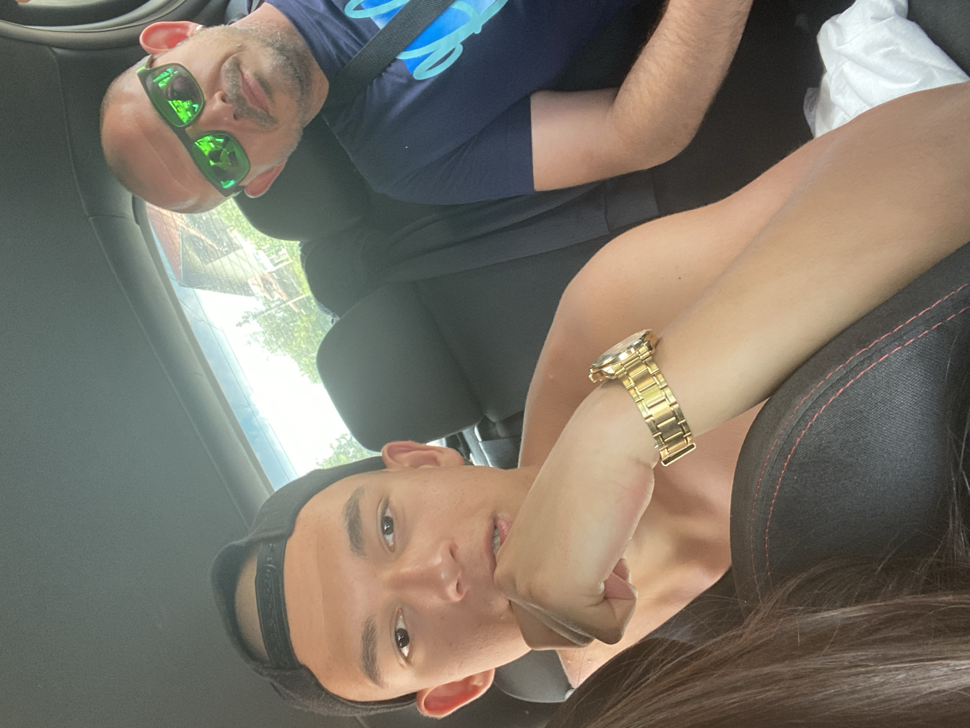 The image shows two men and part of a woman sitting in the back seat of a car. The man in the foreground, wearing a black baseball cap, is looking at the camera with his hand on his chin. He is shirtless and wearing a gold watch. To his right, the second man is wearing sunglasses and a dark blue t-shirt. He is looking towards the camera. A white cloth is visible in the bottom right corner of the image. The background suggests an outdoor environment, possibly with buildings and trees visible through the car window, but there are no specific visual cues to place the location in Sennik, Bulgaria.