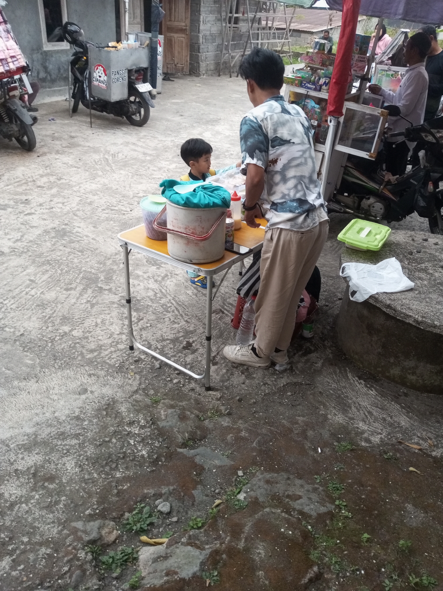 In the city of Kalibeber, Indonesia, a scene unfolds involving a street food vendor and customers. A young boy, wearing a yellow shirt, sits at a low, foldable table, his gaze directed downwards. Beside him, an adult male, dressed in a patterned shirt and beige pants, stands and appears to be interacting with the boy or attending to the stall. On the table, a white bucket with a blue cloth cover sits prominently, alongside a light green container and a bottle with a red cap. A foldable table is set up on a concrete ground, with various items scattered around. In the background, a food cart is mounted on a motorcycle, displaying signs that read "PANGSIT GORENG" and "CIPANG". Another motorcycle is parked to the left of the cart. The environment appears to be an outdoor area, possibly a street or a yard, with a weathered concrete ground and some vegetation. Other individuals are visible in the background, suggesting a public or semi-public space where transactions might be occurring. A white plastic bag and a green plastic container are placed on a large concrete object to the right of the table.