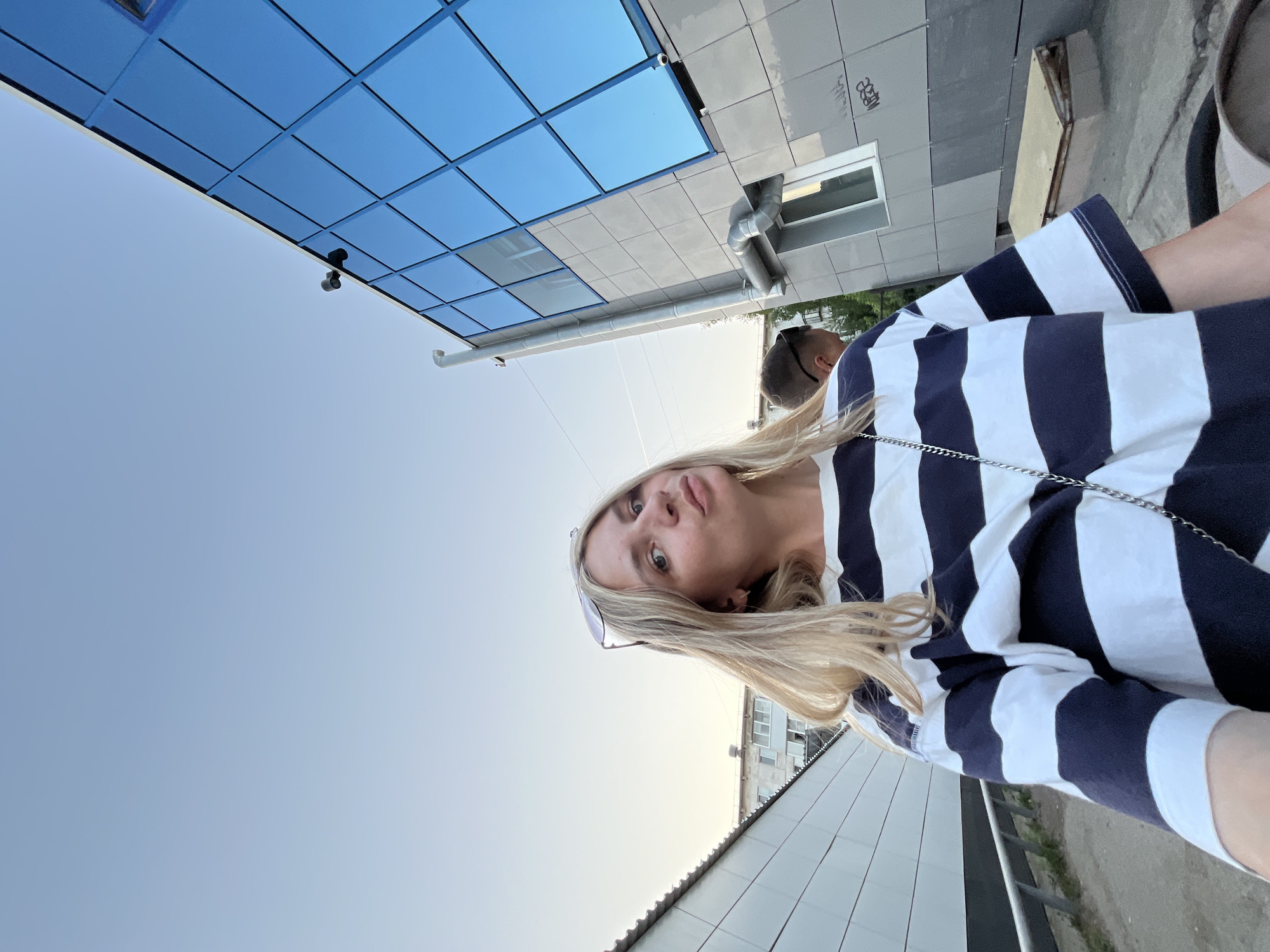 A woman stands outside a building in Chaikovsky, Russia. The building has a blue mirrored facade and gray walls. She wears a striped shirt, and a person stands behind her. The sky is pale blue.