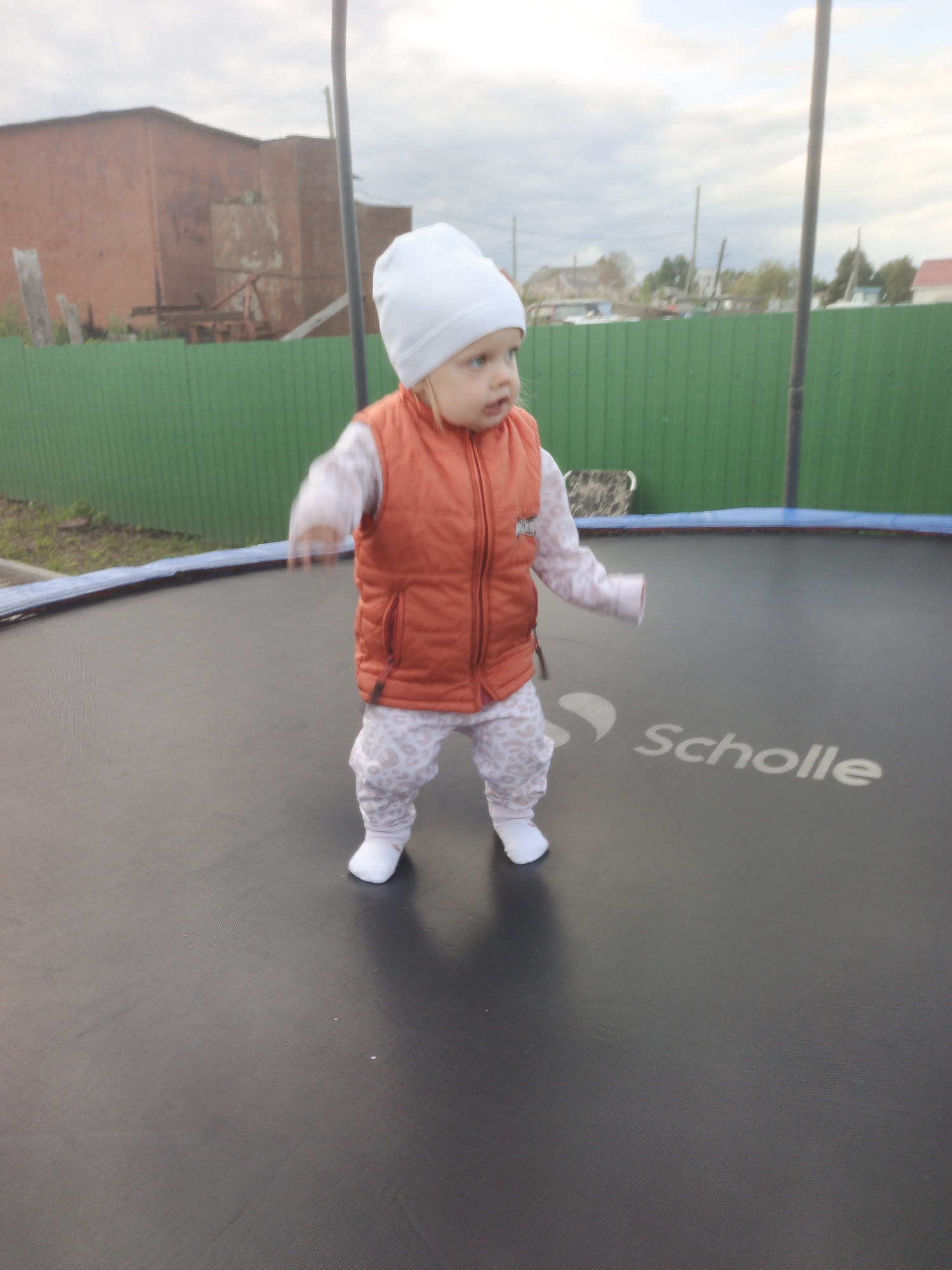 A toddler wearing a white hat and an orange vest over a patterned long-sleeved shirt and pants stands on a black trampoline. The trampoline has the word "Scholle" printed on it in white lettering. In the background, there is a green fence, a brick building, and some trees. The sky is overcast. This scene is located in Medvedyovka, Russia.