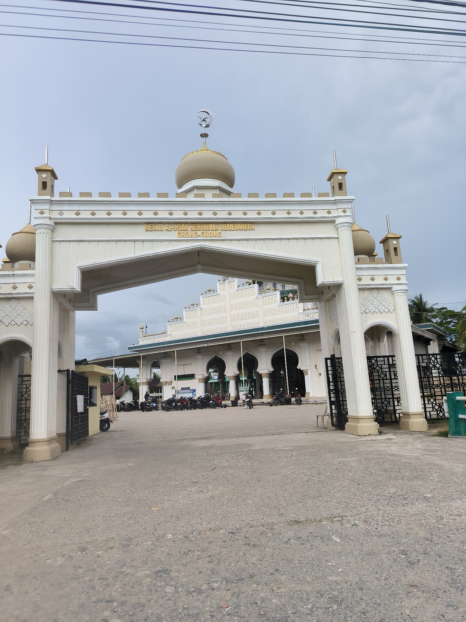 The image captures the grand entrance of the Mesjid Al-Ihsan Kemukiman Meuneubuk Grong-Grong in Grong-Grong, Indonesia. A prominent, ornate cream-colored archway forms the main gateway, featuring decorative pillars, intricate carvings, and small golden-domed minaret-like structures. A central dome on the archway is topped with a crescent moon and star symbol. A golden sign across the arch clearly displays the mosque's name: "MESJID AL-IHSAN KEMUKIMAN MEUNEUBUK GRONG-GRONG".
Beyond the open black metal gates of the archway, the main mosque building is visible, constructed in a similar light-colored style with multiple arched windows and a green trim along its roofline. Several motorbikes are parked in the paved area in front of the mosque, indicating visitors or congregants. The foreground shows a gravel pathway leading up to the entrance.
The scene is set outdoors during the daytime, under an overcast sky. Power lines are visible stretching across the top of the frame. Palm trees and other greenery can be seen in the background behind the mosque complex. No individuals are distinctly visible, but the parked motorbikes suggest a regular day of activity at the mosque.