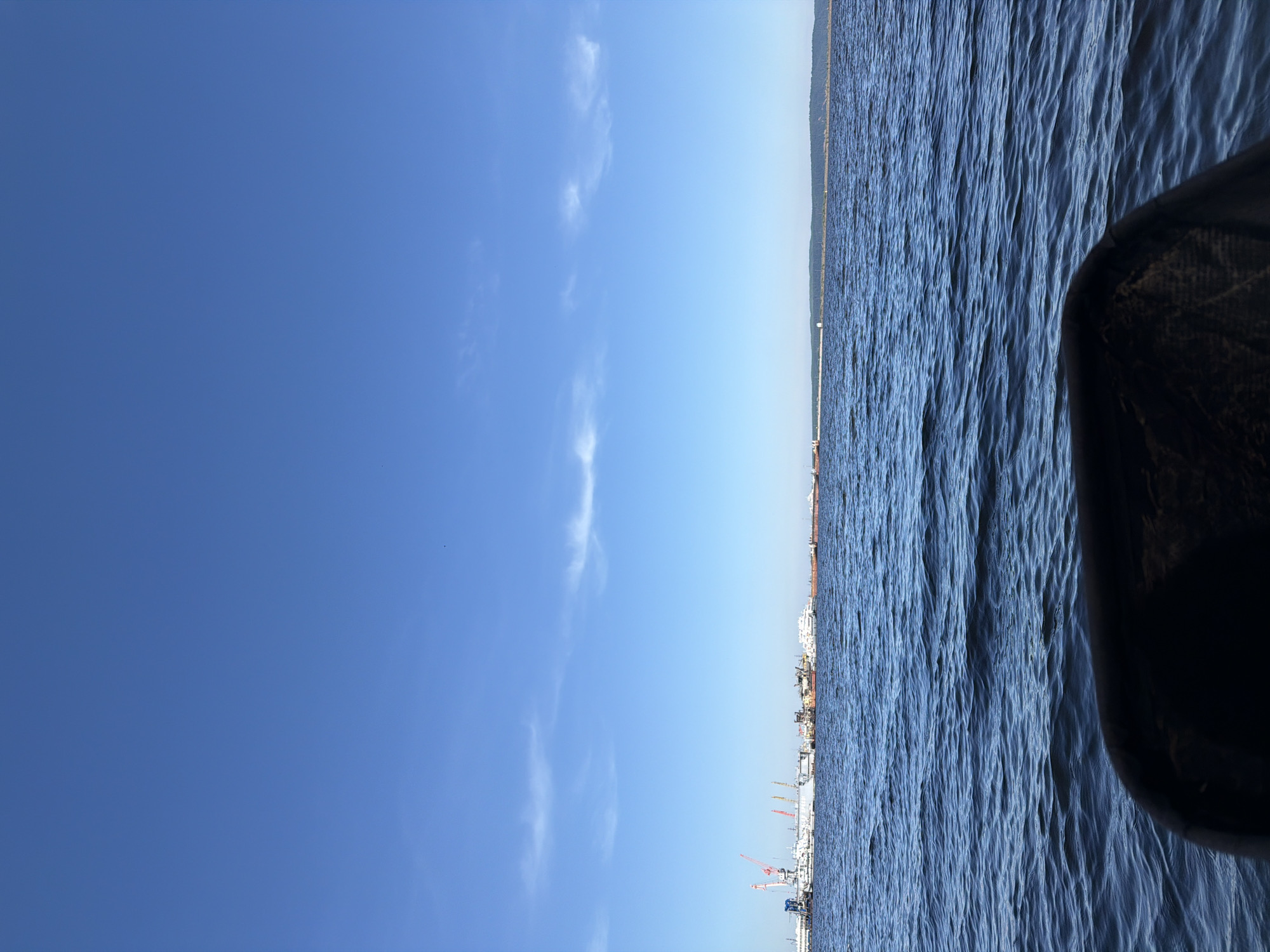 A wide, panoramic shot captures a vast body of water under a clear blue sky. The image is taken from a low vantage point, possibly from a boat or shoreline structure, indicated by a dark, indistinct object partially visible in the bottom right foreground.
The upper portion of the frame is dominated by a bright, clear azure sky, featuring a few wispy, scattered white clouds. The lower half displays a wide expanse of deep blue water, textured with numerous small ripples, suggesting gentle movement or current.
Along the distant horizon, a long, low-lying landmass separates the sky and water. To the left, this shoreline appears urban or industrial, with several cranes (some red, some white) and various ships or barges visible, indicating a port or dock area. Towards the right, the distant land appears more natural, possibly a forested or hilly area.
The scene depicts a calm daytime setting, characterized by bright sunlight and fair weather. No people or discernible text are visible in the frame. The industrial elements on the distant shore, combined with the expansive water body, are consistent with the location context of Chaikovsky, Russia, which is situated on a large river.