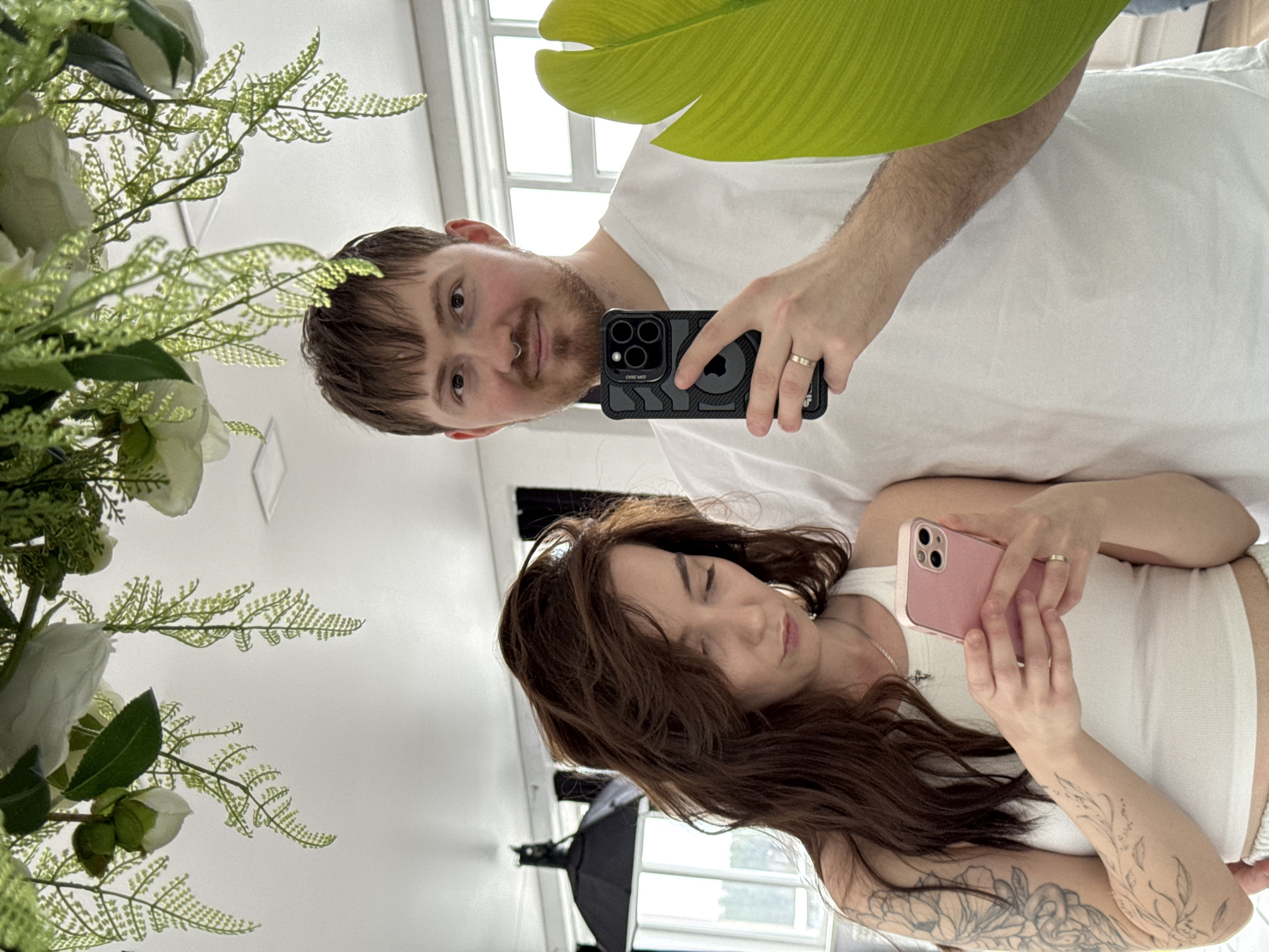 A couple is taking a selfie in a bright, indoor location. The man, positioned at the top of the frame, has brown hair, a mustache, and a nose ring. He is holding a black smartphone with a visible case. The woman, slightly below him, has long, brown hair and is holding a pink smartphone. Both are wearing white tops. The background includes a white wall, a window, and a large green plant to the left. There also appears to be another green plant at the top right. The scene is a reflection, with a lighting setup and another window visible behind the woman. There is no visible evidence in the image to suggest the location is Chaikovsky, Russia.
