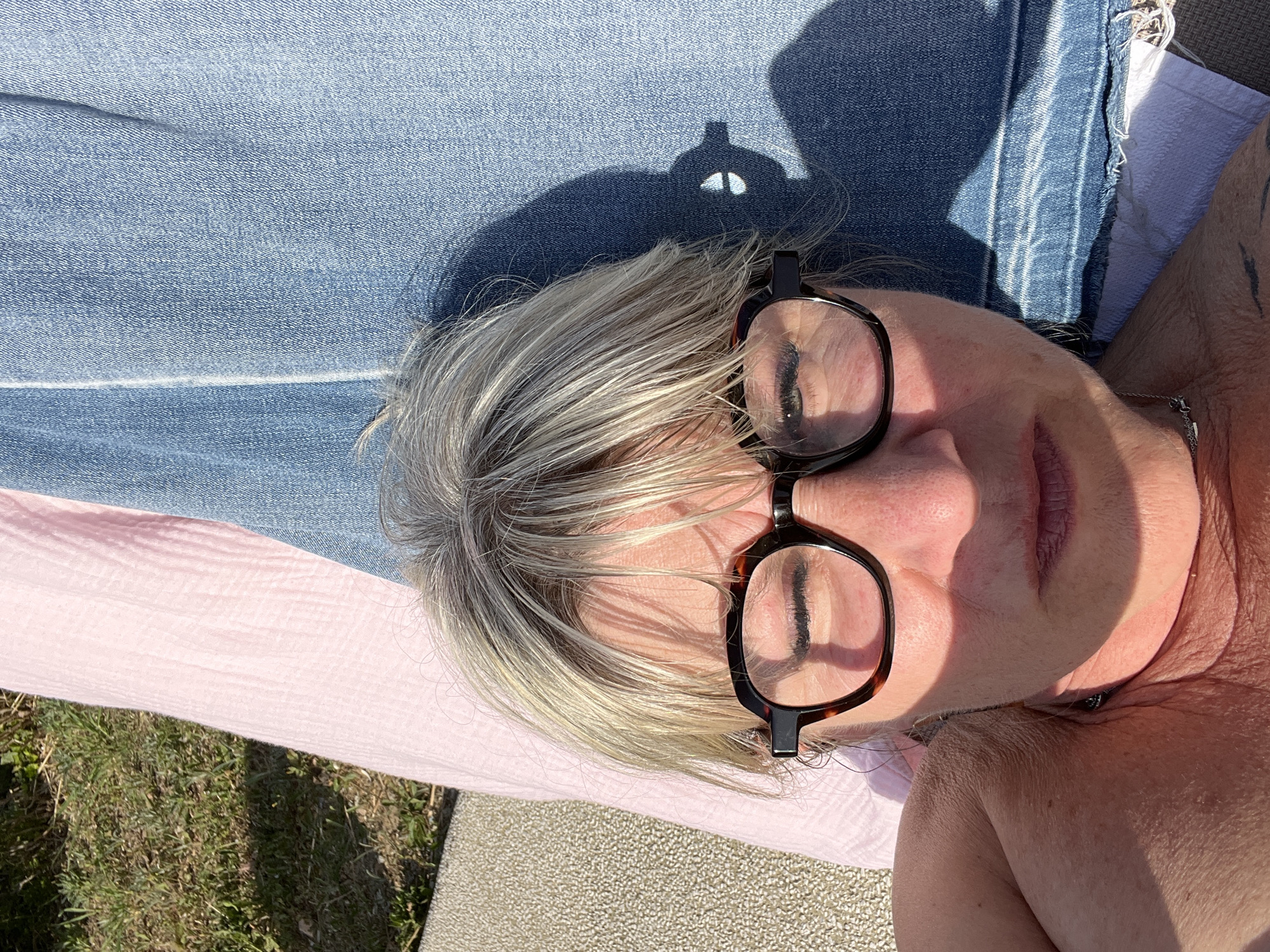A person with gray hair and glasses is lying down outdoors, likely in a backyard or patio setting. The person is positioned on a light pink blanket or cloth, with denim material above. The background shows a patch of grass and a light-colored surface. The presence of sunlight and the clothing style suggest a casual, relaxed setting. There is no definitive information to pinpoint the location as Grimaud, France.