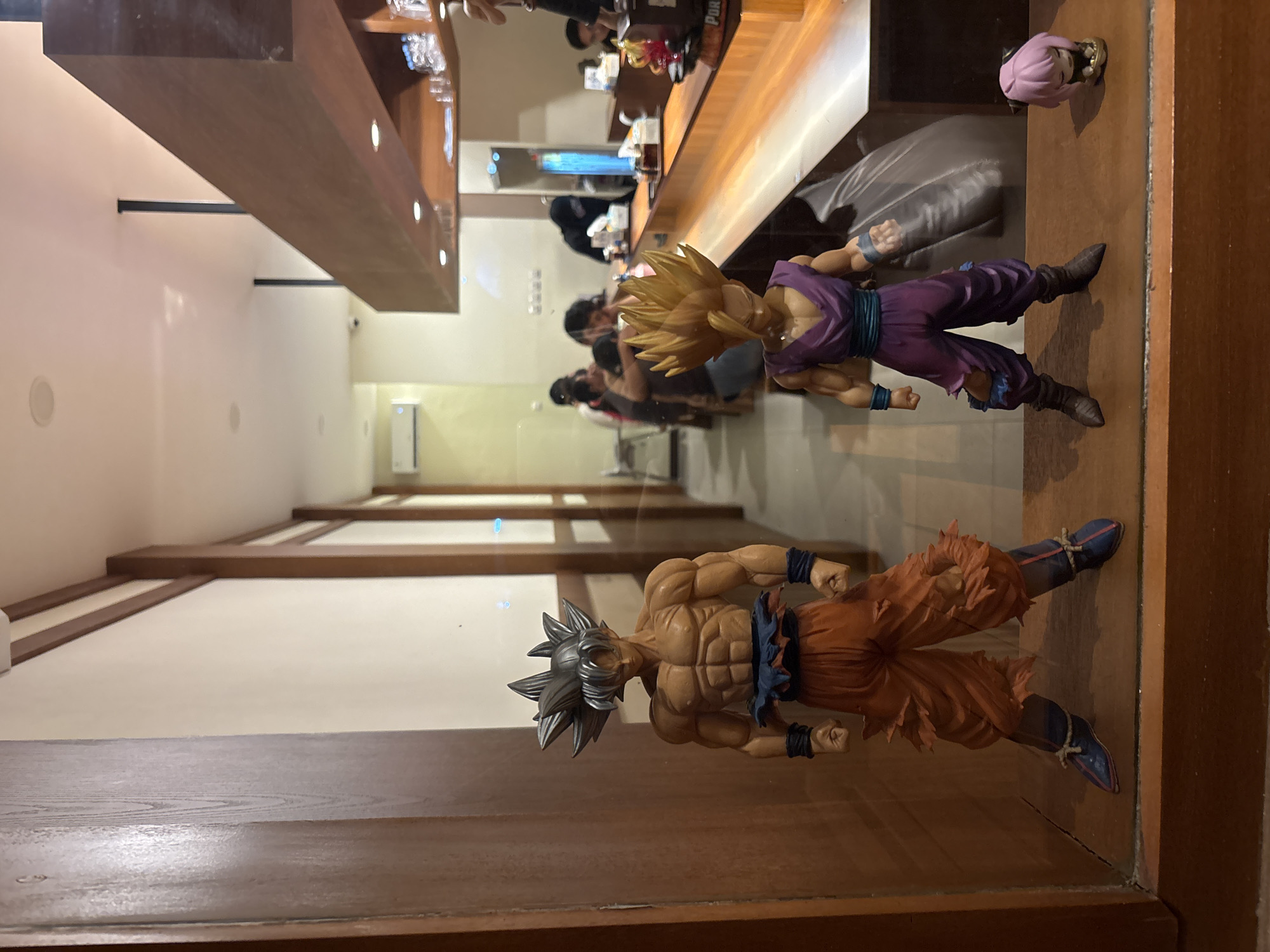 The media file displays two action figures in what appears to be an indoor setting, possibly a restaurant or shop with a counter. The figures are positioned on a wooden surface, with a reflective background suggesting a display case or mirrored wall.
The first action figure, in the foreground, is a muscular male character with spiky dark hair, wearing an orange outfit with a blue sash and dark blue wristbands and boots. The character's pose is dynamic, with fists clenched and a determined expression. This figure is likely representing Goku from the Dragon Ball series in his Ultra Instinct form.
The second action figure, positioned further back and slightly higher, is a male character with spiky blonde hair, wearing a purple gi and blue pants. This figure is also in an action pose. This likely represents Gohan from the Dragon Ball series. A smaller, pink-haired figurine is also visible on the far right.
The background shows a reflection of a space with people, a counter, and shelves with items that are out of focus. The lighting suggests it is daytime or the interior is well-lit. The overall scene appears to be a display of collectible figures in a commercial establishment. There is no discernible text visible in the media file. The location context is West Jakarta, Indonesia, but this is not visually evident from the image itself.
