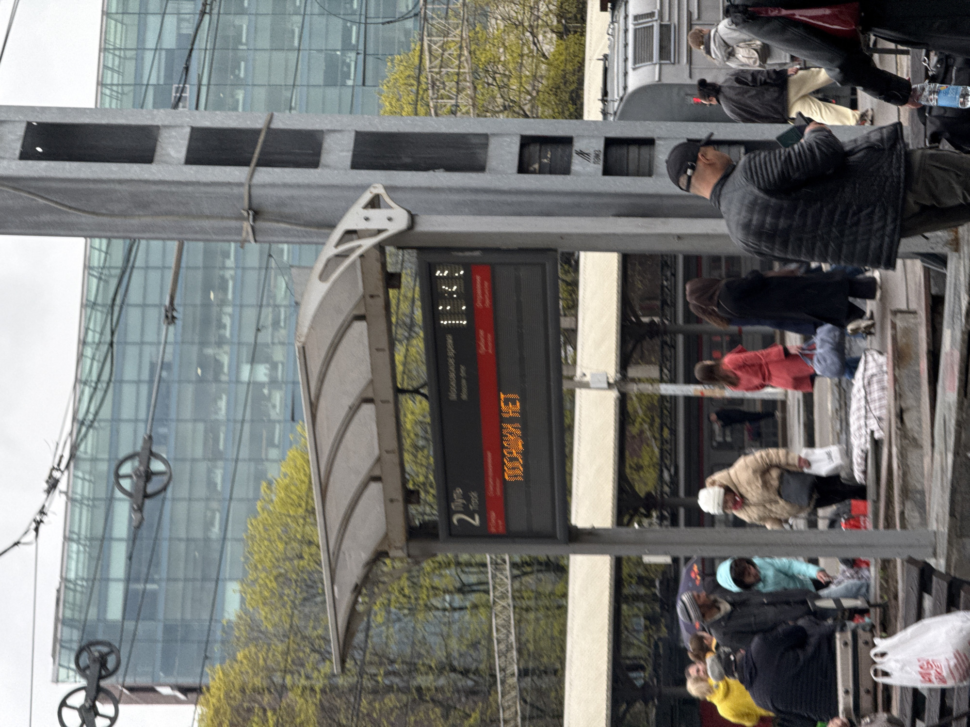 Here is a detailed description of the image:
**Scene:**
The image depicts a busy train platform. There are numerous people walking, standing, or possibly boarding a train.
**Subjects and People:**
- Multiple pedestrians are visible, wearing a variety of clothing, ranging from winter coats to lighter jackets, suggesting variable weather conditions.
- The people are of mixed ages and demographics.
- There are hints of luggage, plastic bags, and other personal items, indicating that people are likely traveling.
**Objects and Details:**
- The image features a train arrival information display screen showing the text "ПОСАДКИ НЕТ" on platform 2, which translates to "No boarding" or "No landing."
- There's a gray metal structure above the platform.
- In the background, a large glass-clad building, possibly an office or commercial structure, can be seen, suggesting an urban setting.
- There are wheels and cables in the background, possibly part of the infrastructure of the train system.
- The photograph is taken at eye level from an urban public train station.