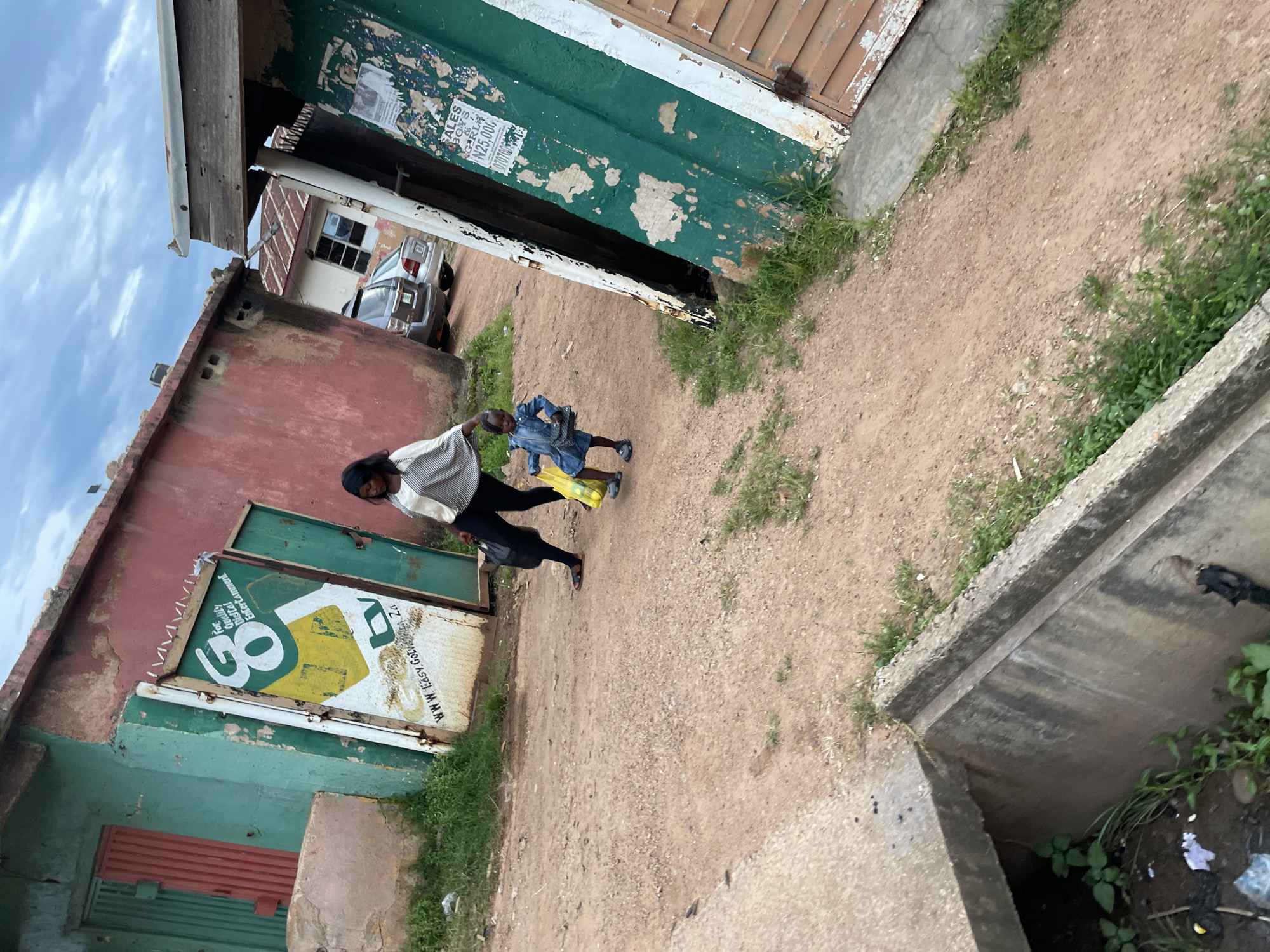 In Jos, Nigeria, on a cloudy day, a woman and a child walk across a dirt path lined with weathered buildings. The woman, wearing a striped top and dark pants, carries a black bag. The child, dressed in a denim outfit, holds two yellow bags. One building, painted red and green, has a partially opened metal gate with a "GO" logo and text that appears to be an advertisement. Another building, also green, displays a poster with text including "SALES BOYS & GRLS" and "N25,000". Two cars are parked in the background. A concrete drainage ditch, filled with some water and debris, is visible in the foreground.