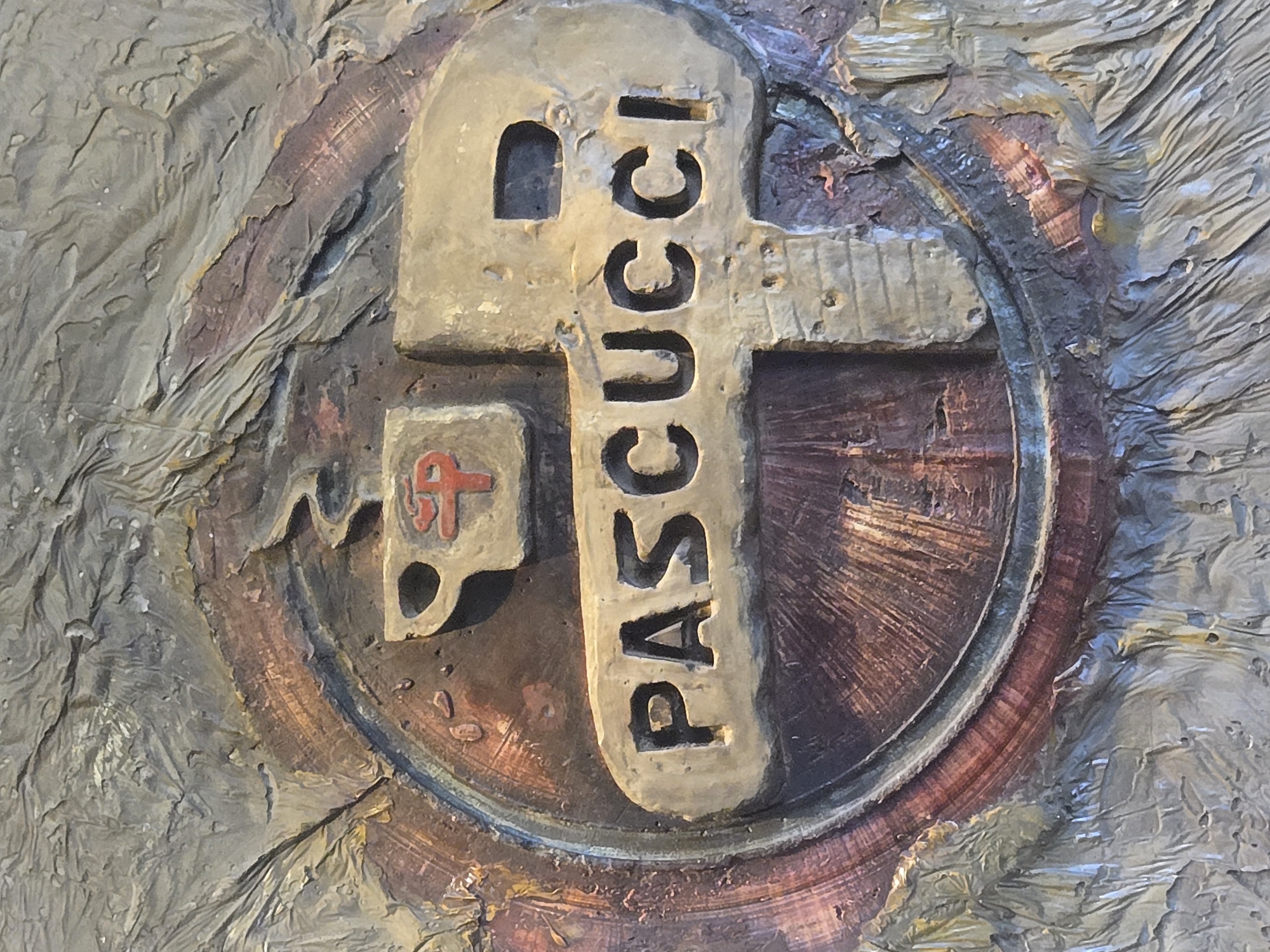 This image shows a close-up of a plaque or relief with the name "PASCUCCI" inscribed vertically on a key shape. To the left of the key is a smaller, cup-shaped emblem bearing a red symbol resembling a cross or stylized initial. The key and emblem are set within a circular frame that appears to be metallic, possibly bronze or brass with an aged patina. The background is a textured, possibly stucco-like surface.
This plaque is located in the Stari Bar of Bar, Montenegro.