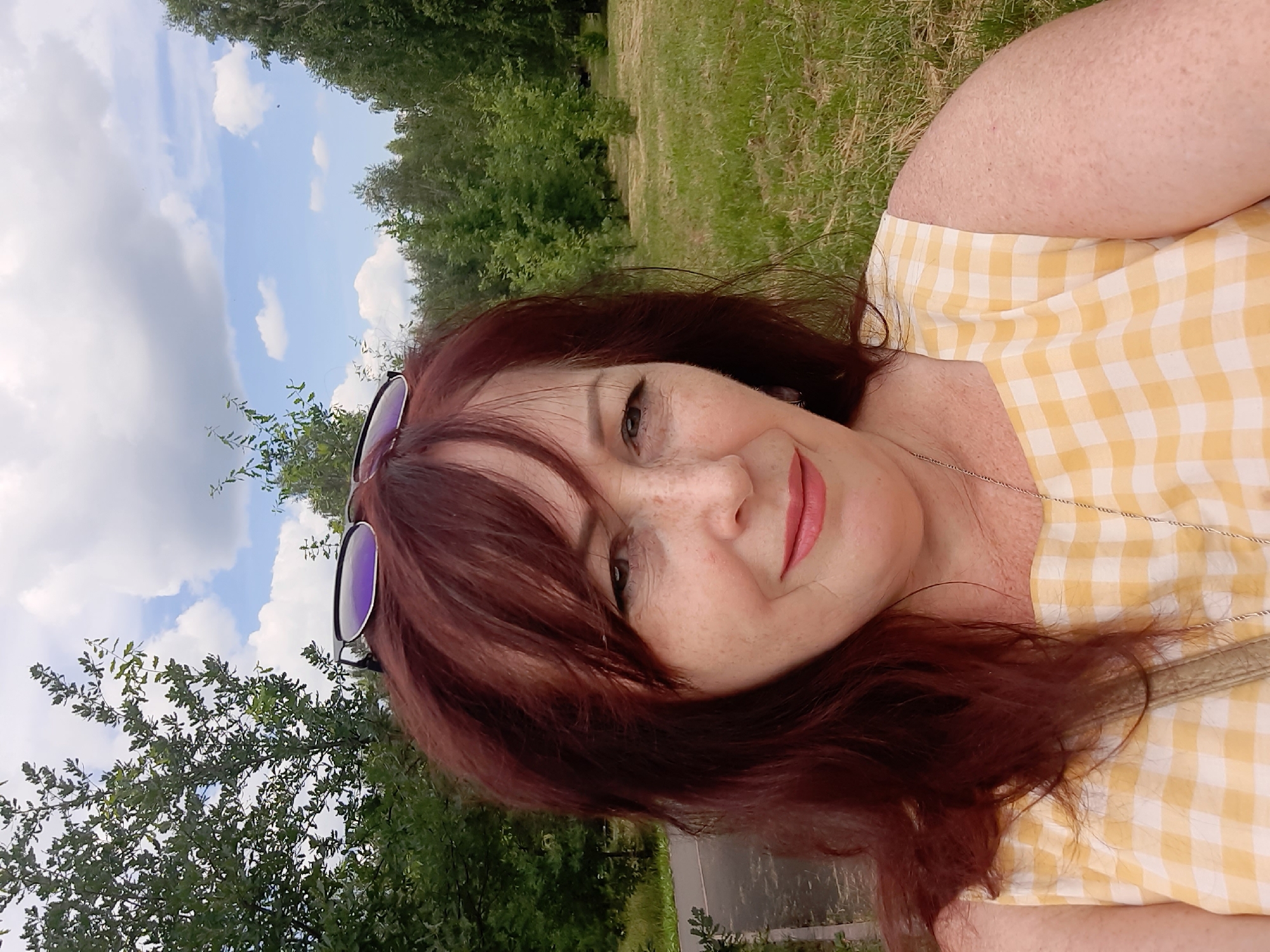 A woman with auburn hair and freckles, wearing a yellow and white checkered dress and sunglasses on her head, takes a selfie in a park. The background consists of trees, a paved path, and a cloudy sky. The image appears to be taken outdoors in a natural setting.