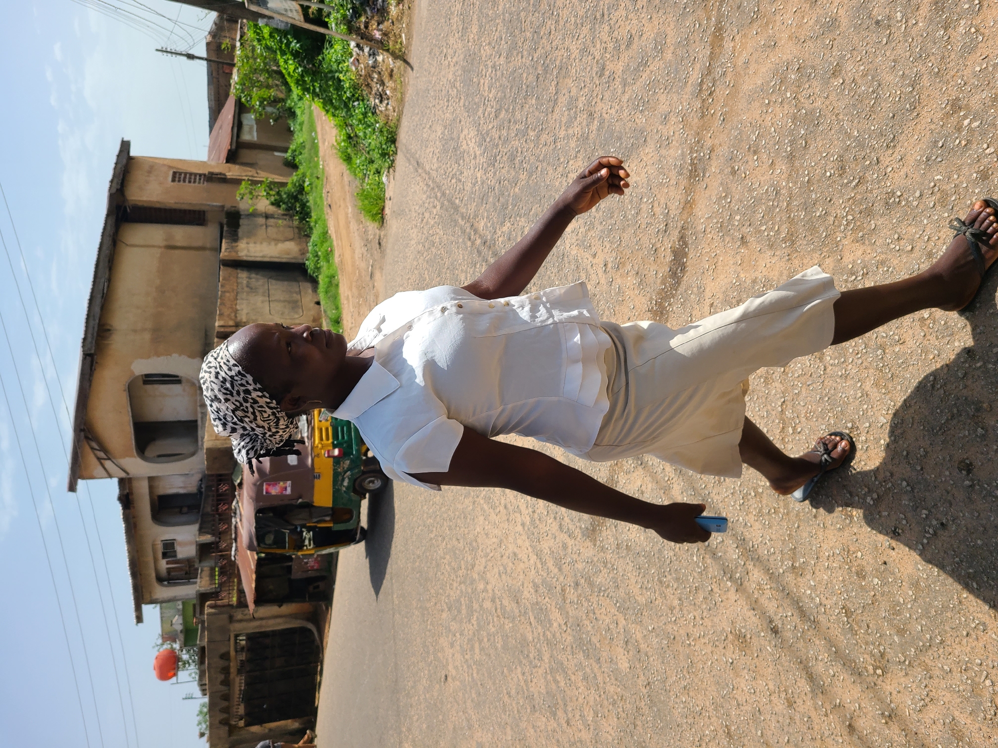 An outdoor, eye-level, medium shot captures a Nigerian woman walking across a dusty, light-colored road in Jos, Nigeria. The woman, who appears to be of African descent, is wearing a white button-down shirt with ruffled details at the waist and a light-colored skirt that reaches her knees. She has a patterned headscarf covering her hair and is looking off to the right. In her right hand, she holds a blue mobile phone. Her shadow is cast across the road to her left.
In the background, a yellow and green auto-rickshaw, also known as a "Keke Napep," is parked on the side of the road, partially obscured by the woman. Behind the Keke is a building with a weathered, tan exterior and a corrugated metal roof. Further back, another building with similar architecture is visible, along with some green foliage and power lines. The sky is clear and blue with some faint clouds. The overall environment suggests a moderately urban or semi-urban setting in Jos, Nigeria.