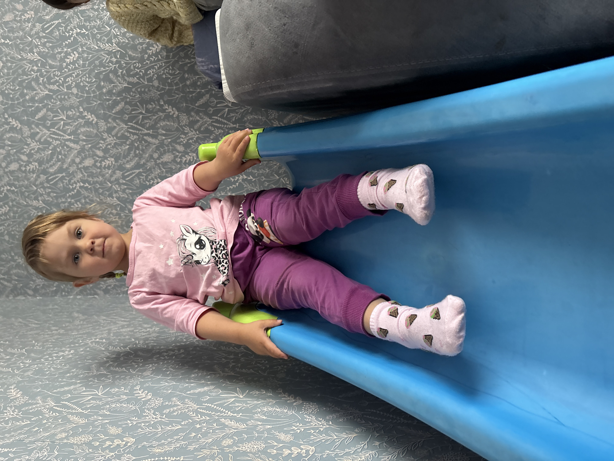 The scene depicts an indoor residential setting in the city of Фоки, Russia. A young child, approximately 2-4 years old, with light-colored hair styled into two small braids secured with green elastics, blue eyes, and fair skin, is seated upright on a vibrant blue plastic children's slide. The child is wearing a pink long-sleeved t-shirt featuring a graphic of a white pony with black and white accents, purple sweatpants, and light pink ankle socks adorned with small brown and green cake slice patterns. Both of the child's hands are gripping the light green plastic handles situated on either side of the slide's upper section. The child's feet are placed flat on the blue sliding surface, facing directly towards the observer with a neutral expression.
To the left of the child, a wall is covered with light blue wallpaper featuring a repeating pattern of intricate white line-art botanical elements, including various leaves and small flowers. To the right of the child, a portion of a dark grey or blue upholstered sofa or armchair is visible. In the upper right corner, a segment of another individual is discernible, wearing a light yellow or cream-colored knit sweater.