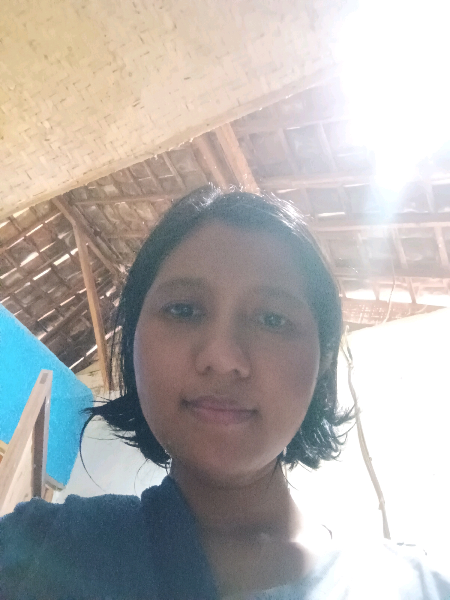The image is a selfie taken from a low angle, focusing on the face of a young person with dark, shoulder-length hair. The person is wearing a dark-colored top. The background shows a thatched ceiling and wooden beams, suggesting an indoor location, possibly a traditional dwelling. To the left, there is a bright blue wall with a wooden structure attached. The lighting is strong, with a significant glare on the right side of the image, indicating a bright light source, possibly the sun. The specific location in Canga'an, Indonesia cannot be determined from the visual information in the image.