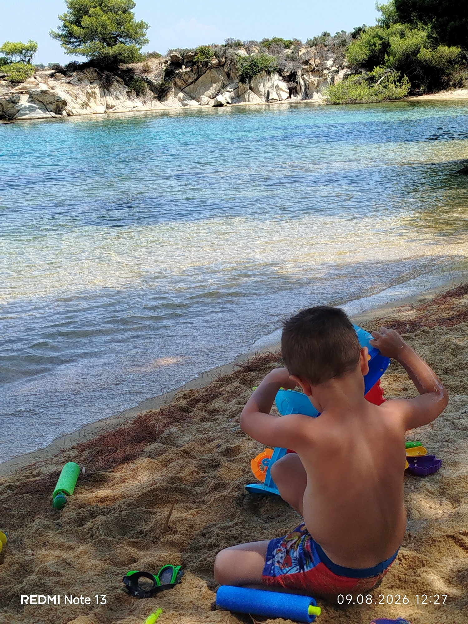 A young boy plays with sand toys on a sandy beach in Sithonia Municipal Unit, Greece. The boy is seen from behind, wearing colorful swimming trunks and playing with a blue and red toy bucket. Various other beach toys, including goggles and a green water bottle, are scattered in the sand around him. The clear turquoise water of the Aegean Sea gently laps the shore in the background, with rocky cliffs and lush greenery visible along the coastline under a bright, sunny sky. The timestamp "09.08.2026 12:27" is visible in the lower right corner of the image, along with the text "REDMI Note 13".