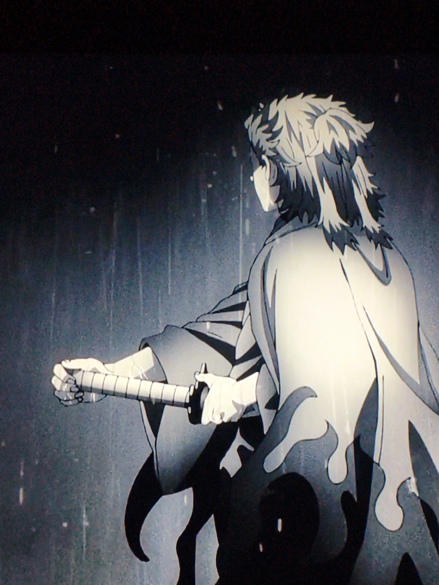 The image shows an anime character from the side, holding a sword. The character has spiky, blonde hair and is wearing a white haori with flame-like patterns. The background is dark and appears to be raining. There is no visible indication of the city of Vladivostok, Russia, in this image. The scene is static, with the character posed in a ready stance. The lighting is dim, and the overall tone is dramatic.