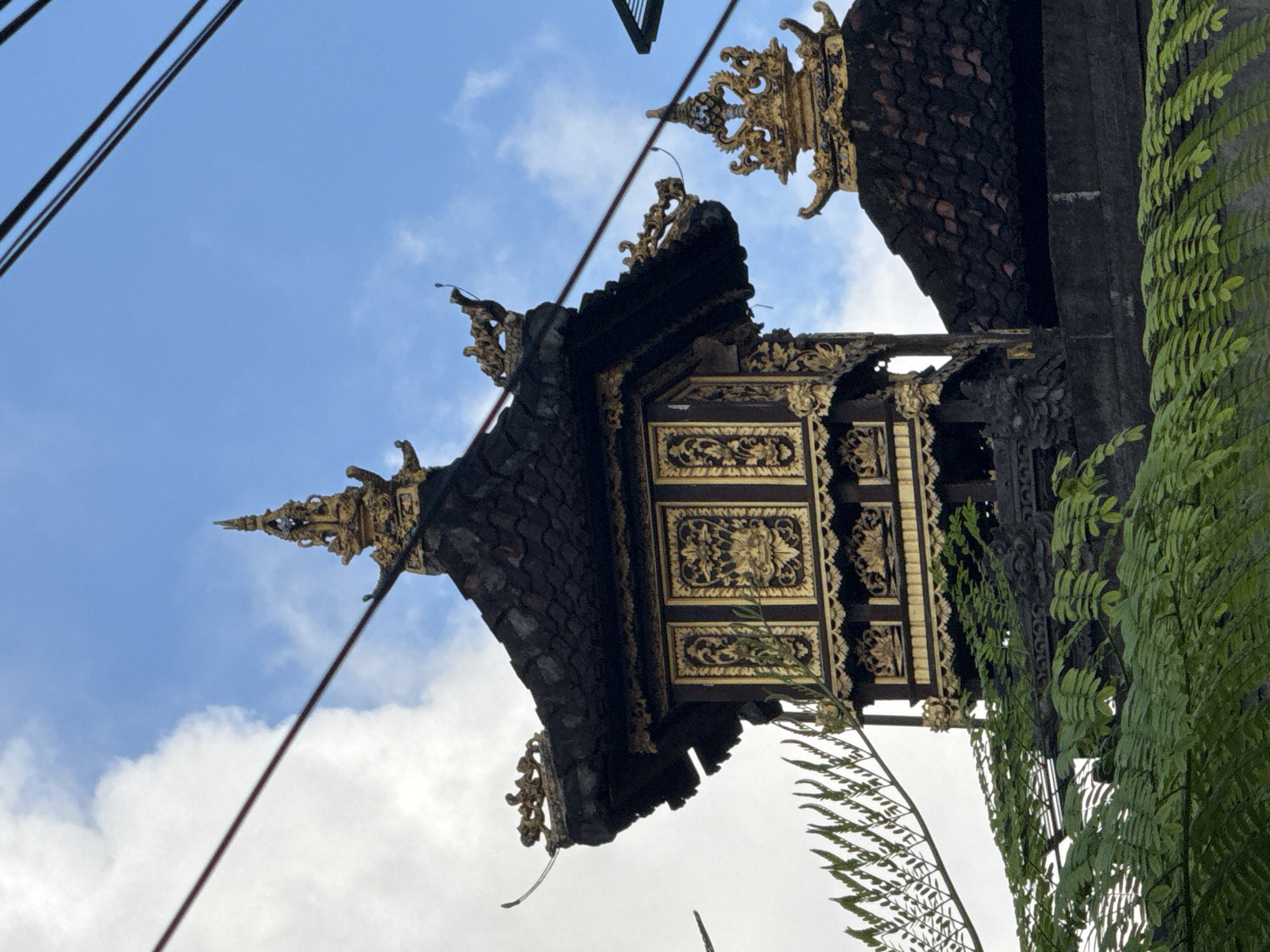 The image depicts a section of Balinese architecture against a backdrop of a partially cloudy blue sky. The building is adorned with intricate gold detailing. The lower right portion of the image features green foliage, possibly vines or ferns, growing against the side of the structure. Electrical wires run across the top left of the image. The location is likely Kuta, Indonesia, based on the architectural style.