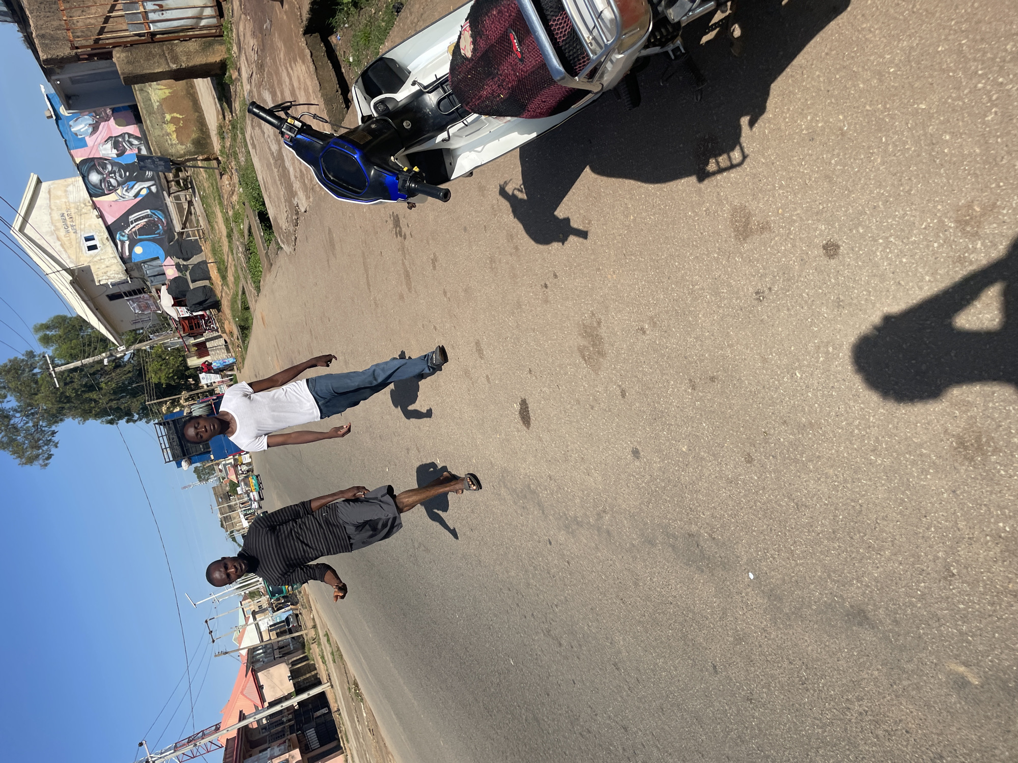 A street scene in Jos, Nigeria, features two young Black men walking on an asphalt road under a clear blue sky. The man on the left wears a black and gray striped long-sleeved shirt and dark gray shorts. He is looking towards the viewer and has his right hand outstretched, palm open, as if offering something or gesturing. He is wearing dark flip-flops. The man on the right wears a white t-shirt and blue jeans. He is looking ahead and slightly to his left.
To the right of the men, a white scooter with blue accents is parked on the side of the road. The scooter has a black and red seat cover with a mesh overlay. The road surface shows some oil stains and debris. In the background, buildings line the street, one of which features vibrant murals depicting two women with glasses and colorful drinks, along with text that reads "BULL WOMEN AFFAIRS". Trees and utility poles are also visible, contributing to the urban environment of Jos. Shadows of the men and the scooter are cast on the road.
