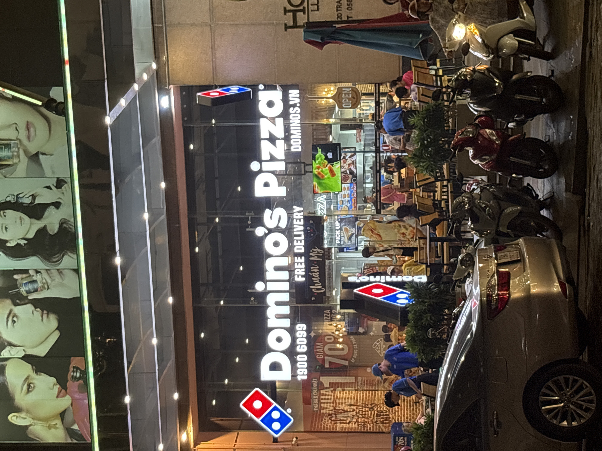 This image depicts the exterior of a Domino's Pizza restaurant in Nha Trang Ward, Vietnam, at night. The brightly lit storefront features the iconic Domino's Pizza logo and signage, including a phone number "1900 6099" and the website "DOMINOS.VN". Inside, the restaurant appears to be busy with customers and staff visible through the large glass windows. A large digital display showing a thermal map of what appears to be a pizza is also prominent.
Outside the restaurant, several motorcycles are parked. A silver sedan is parked on the left side of the frame. The street in front of the establishment is illuminated by the store's lights and streetlights, casting reflections on the wet pavement, suggesting it might have recently rained. The upper portion of the building features large, stylized portraits of people. The overall scene conveys a lively nighttime atmosphere of a popular fast-food establishment.