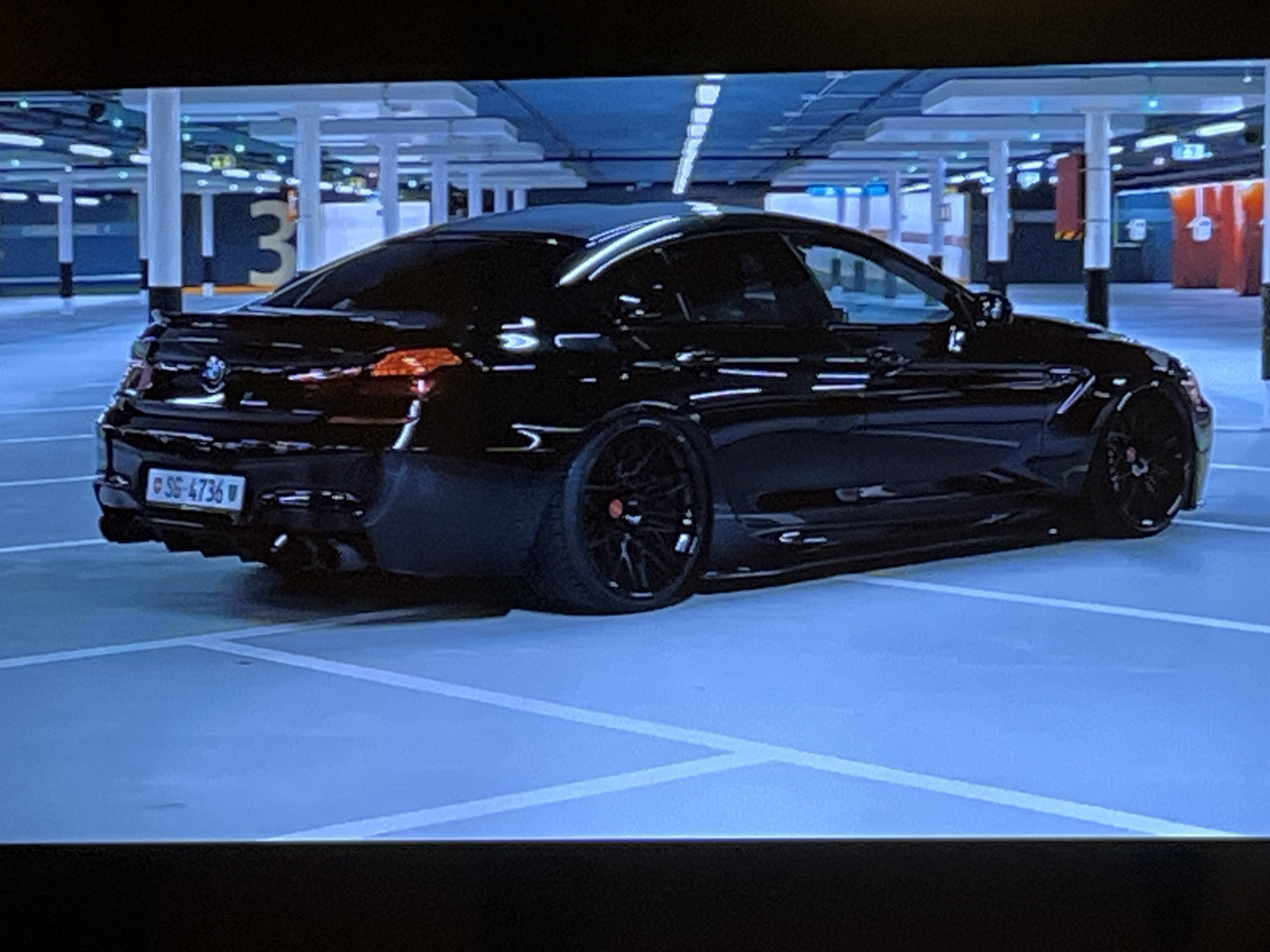The image shows a black sedan parked in a multi-story parking garage. The car is viewed from the rear-left quarter. It has a spoiler and dual exhaust pipes. The license plate is visible and reads "SG-4736 W". The parking garage has white pillars and a blue ceiling with lights. There are signs with the number "3" visible in the background. The floor of the garage is light blue with white lines marking parking spaces. The image does not provide any information about the city of Kharkiv, Ukraine, or any people.