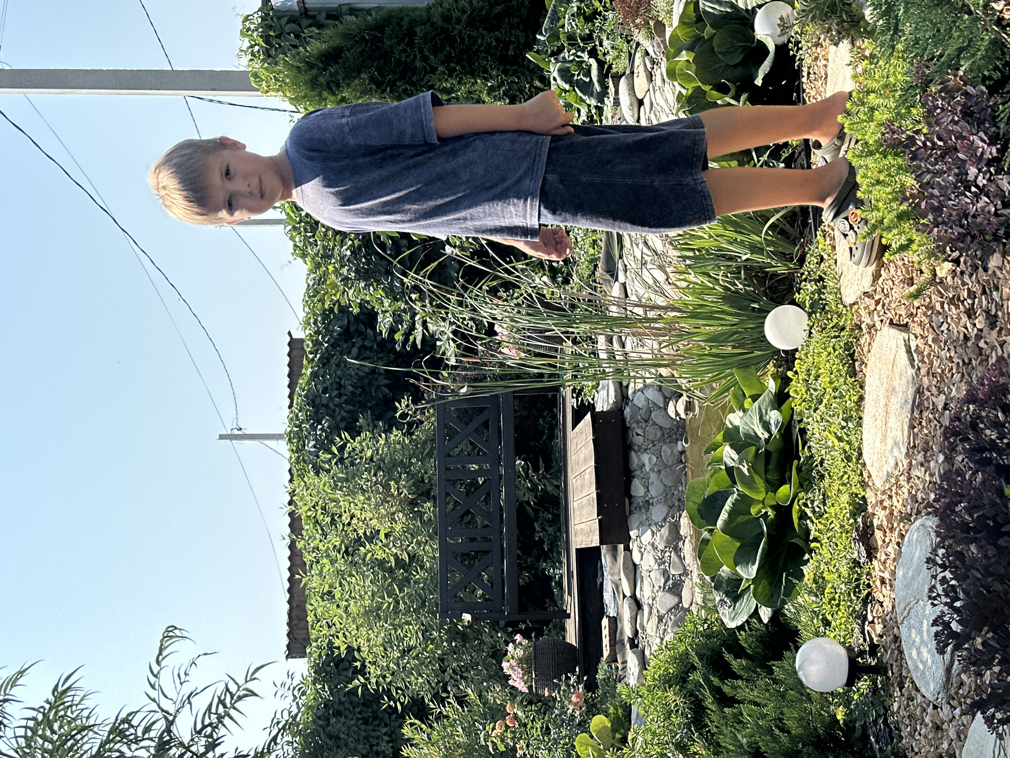 A young boy with blond hair, wearing a dark grey t-shirt and shorts, stands in a garden on a sunny day. He is facing the camera and looking directly at it with a neutral expression. The garden features a small pond with rocks and aquatic plants, a wooden bench, and various greenery, including bushes, trees, and flowers. Two round, white garden lights are visible on the ground. In the background, there is a wooden fence, a roof, and utility poles with wires against a clear blue sky. The location is identified as Samarsky, Russia.