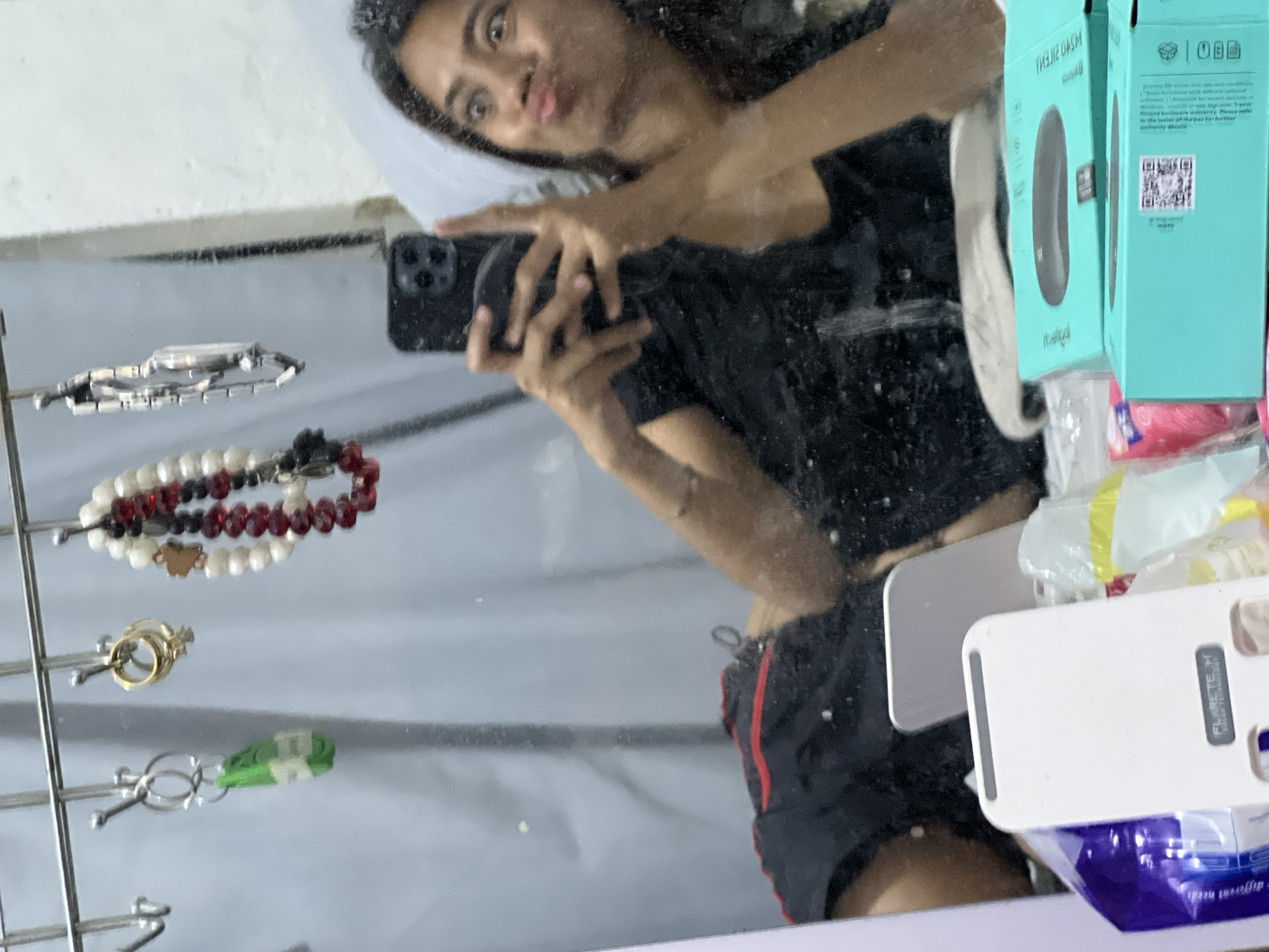 The image is a reflection in a mirror showing a young woman taking a selfie. She is lying down and is wearing a black t-shirt and black shorts with red trim. She is holding a smartphone in her right hand. In the reflection, several items are hanging on hooks above her, including bracelets and rings. Below her, on a surface, are various objects, including a white electronic device with the brand name "FLARETECH" and some teal-colored boxes. The background appears to be a room with light-colored walls and curtains.
This scene is not specific to any particular location in Davao City, Philippines, or any other city. It depicts a common indoor setting, likely a bedroom or dressing area, where a person might take a selfie. Without additional visual cues or context, it is impossible to pinpoint its location.