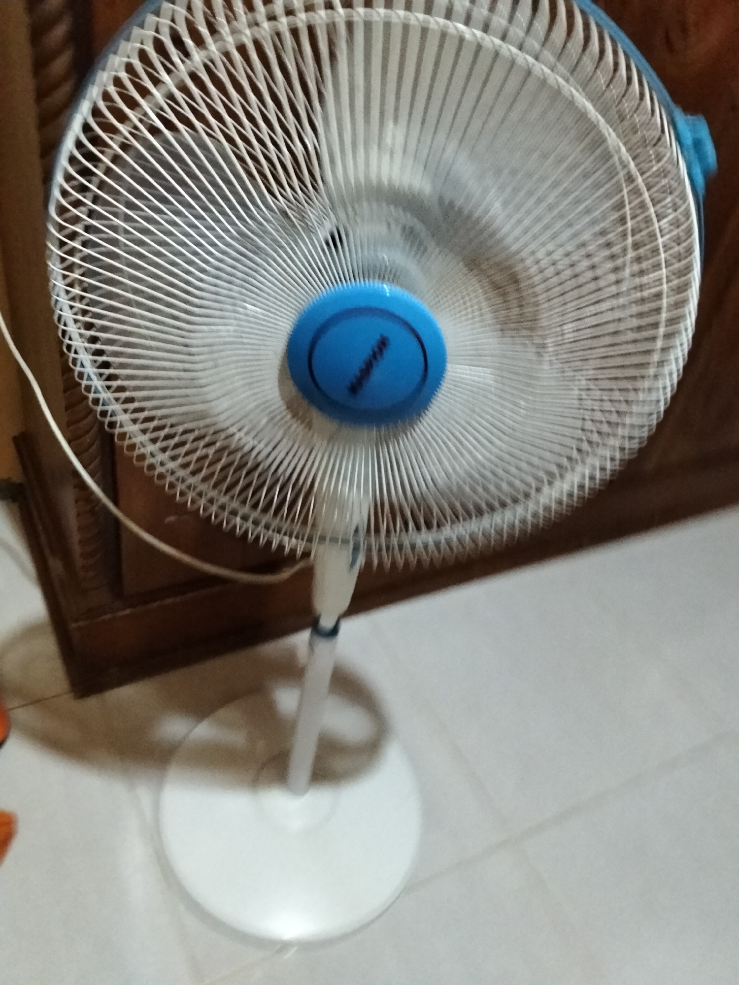 The image shows a white oscillating fan with a blue central hub. The fan is standing on a white circular base. It is positioned in front of a wooden cabinet with carved details. The fan is on and its blades are blurred, indicating motion. The floor is tiled in a light color. The scene is indoors, likely in a residential setting in Payagaboh, Indonesia, based on the provided context. There are no people visible in the image.