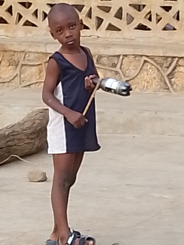 A young, slender boy stands outdoors, holding a stick attached to a plastic bottle. He wears a dark blue and white sleeveless dress with sandals on his feet. His gaze is directed forward, conveying a somewhat serious expression. A wall with a unique patterned design sits behind him. A log rests to his left, with a small rock nearby. The ground is a concrete surface.