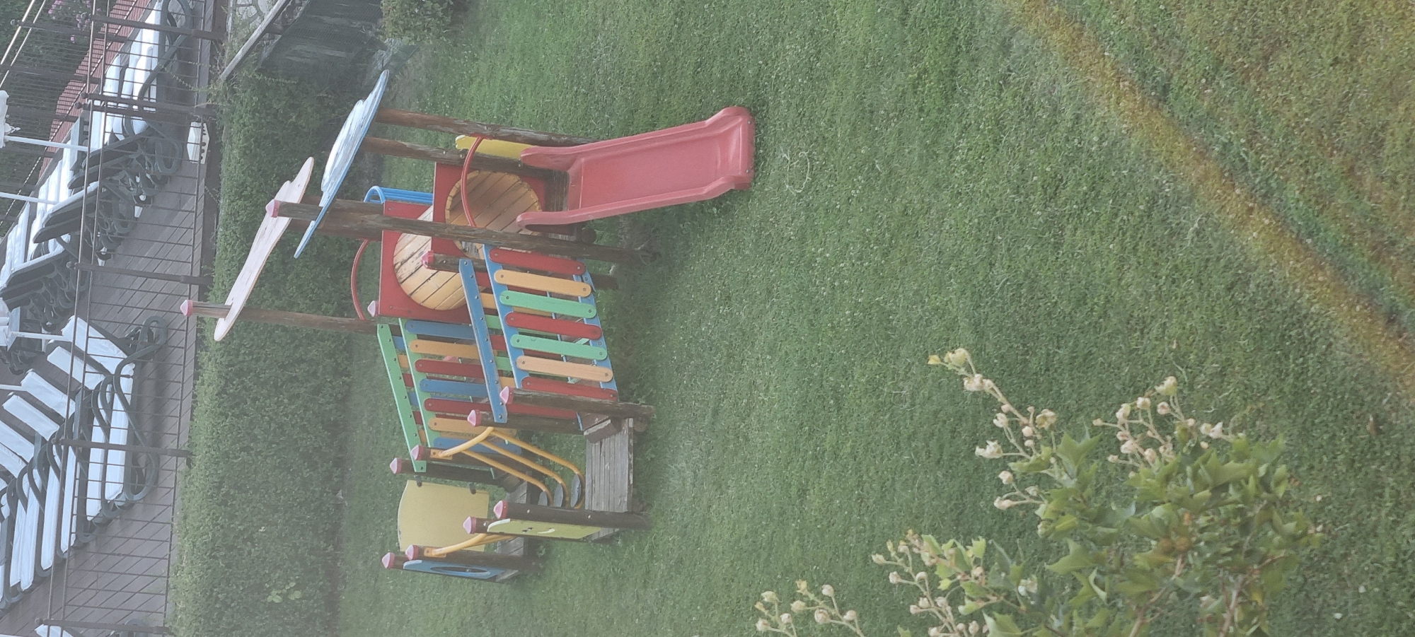The image depicts a brightly colored children's playground structure situated on a grassy area. The playground features multiple sections, including a red slide, a tunnel with a wooden interior, and various platforms and railings. The railings are made of vertical wooden slats in alternating colors of red, blue, green, and yellow. The structure is supported by wooden posts. In the background, there is a dense green hedge, and beyond that, a row of lounge chairs with white cushions, suggesting a resort or hotel environment. The overall setting appears to be outdoors, with a focus on recreational facilities. The location is identified as Sveti Vlas, Bulgaria.