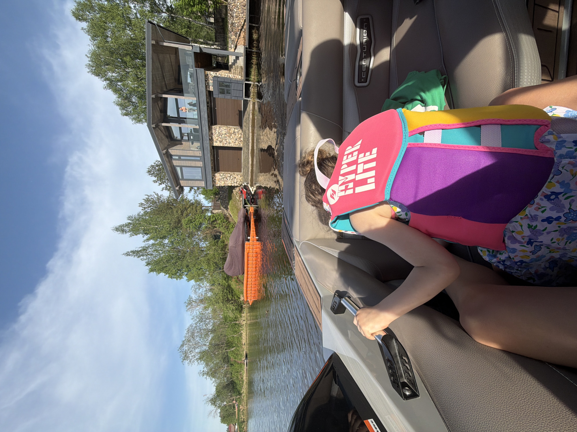 A girl in a colorful life vest is seated in a Centurion boat on a lake. The boat is near a lake house with a boat lift. The location may be Терехово, Russia.