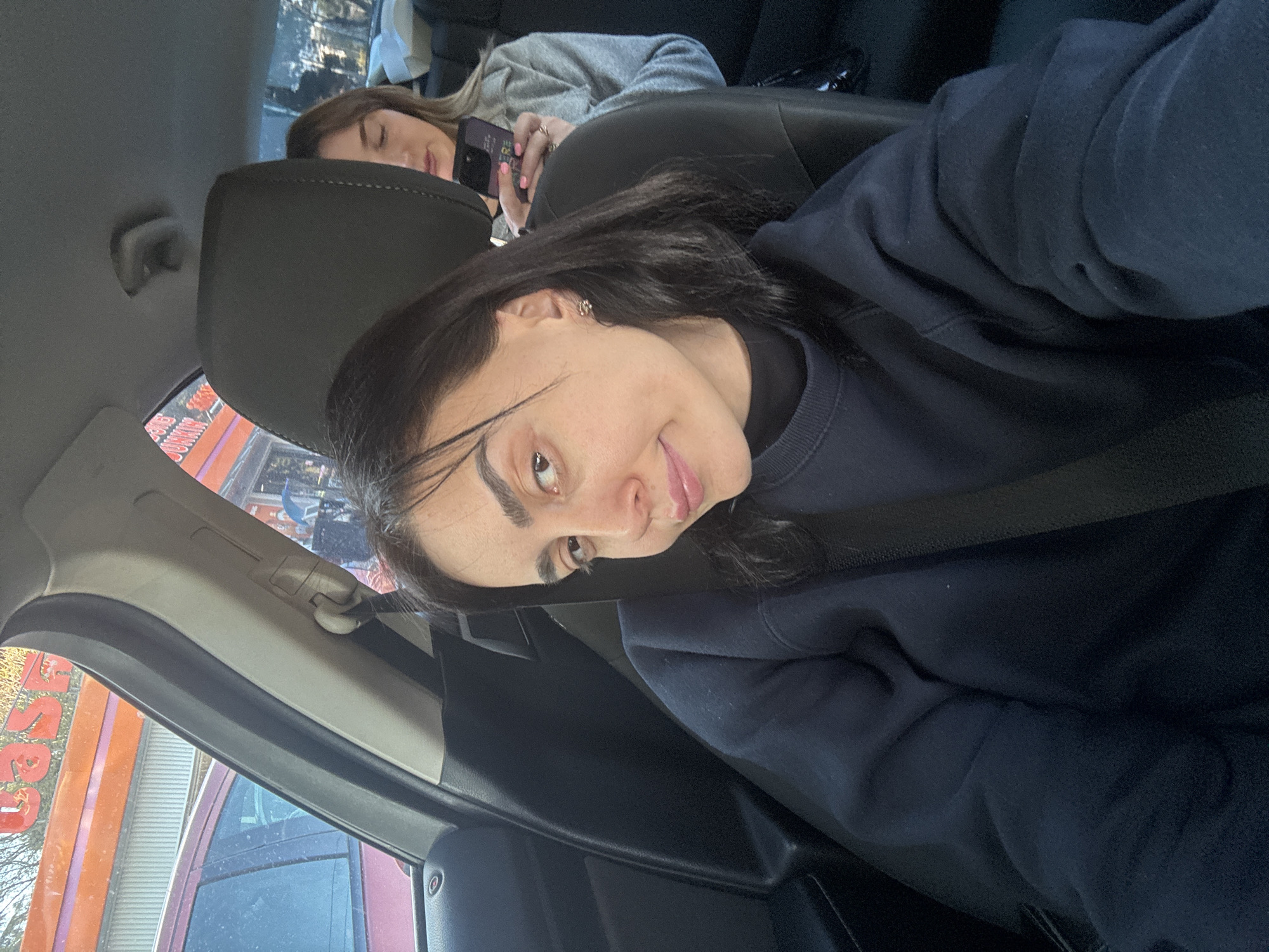 The image shows two people inside a car. The foreground person, presumably the photographer, is wearing a dark blue sweater and a seatbelt. They are facing the camera with a slight smile. Behind the headrest, another person is reclining, seemingly asleep, holding a mobile phone. The environment visible through the car window suggests an urban setting with commercial buildings, specifically displaying visible colored signs and lettering. Given the location details provided, it is possible the scene is in the city of Tbilisi, Georgia.
