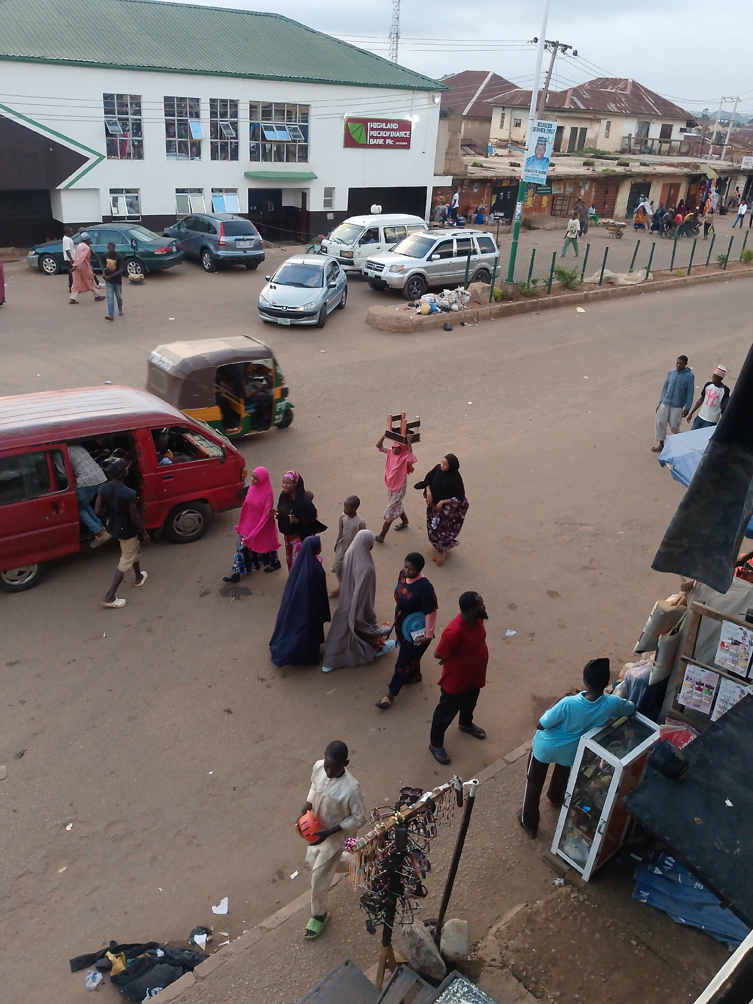 This image captures a bustling street scene in Bukuru, Nigeria. Several people are walking on a paved road and sidewalks, some appearing to be shopping or commuting. Various vehicles are present, including a red van with its doors open, a tuk-tuk, and several cars parked or driving.
A group of individuals is seen near the red van, with some entering or exiting the vehicle. Further down the road, a woman in a pink hijab carries a wooden structure on her head, while others in various traditional attire walk along. Some people are browsing or working at stalls set up along the sidewalk, with displays of goods like eyeglasses and other items visible.
In the background, buildings line the street, some appearing to be businesses. One building has a sign that reads "HIGHLAND MICROFINANCE BANK Plc". Another sign displays a portrait of a man and text that appears to be related to an election or political campaign, with "SELFLESS SERVICE 2027" visible.
The time of day appears to be late afternoon, with the lighting suggesting the sun is not at its highest point. The sky is overcast, indicating potentially cloudy weather. The overall atmosphere seems to be one of everyday activity and commerce in a Nigerian town.