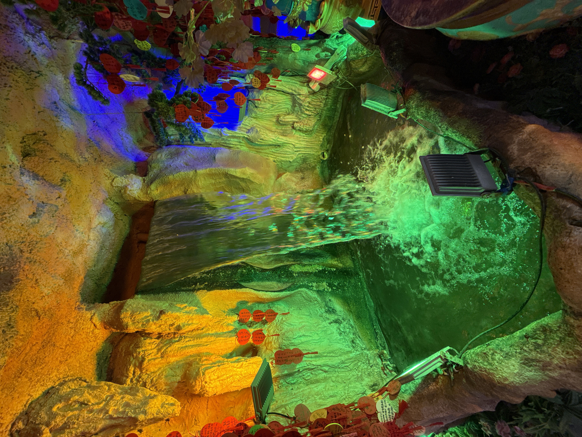 This image depicts a cave-like structure in the Yuzhong District, China. Artificial rock formations are illuminated with a variety of colored lights, including yellow, green, and blue. A stream of water flows through the structure, creating a small waterfall. Several electrical lights are mounted on the rock formations. The scene is decorated with hanging ornaments.