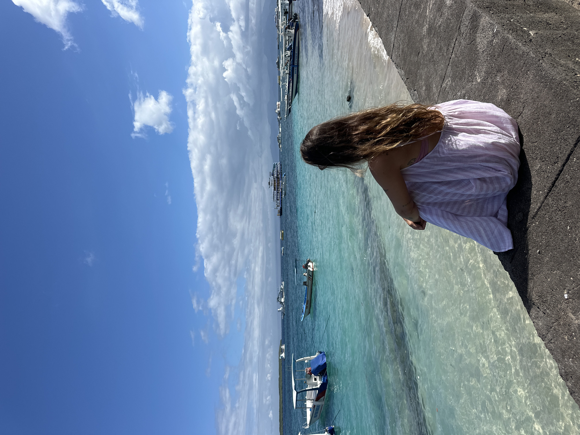 A woman in a light pink and white striped dress is sitting on a dark gray concrete pier next to the clear turquoise water of a beach. Her brown hair is down and partially covers her face as she looks towards the water. Numerous boats are visible in the distance on the water. The sky is blue with scattered white clouds. The location is Nusa Penida, Indonesia.
