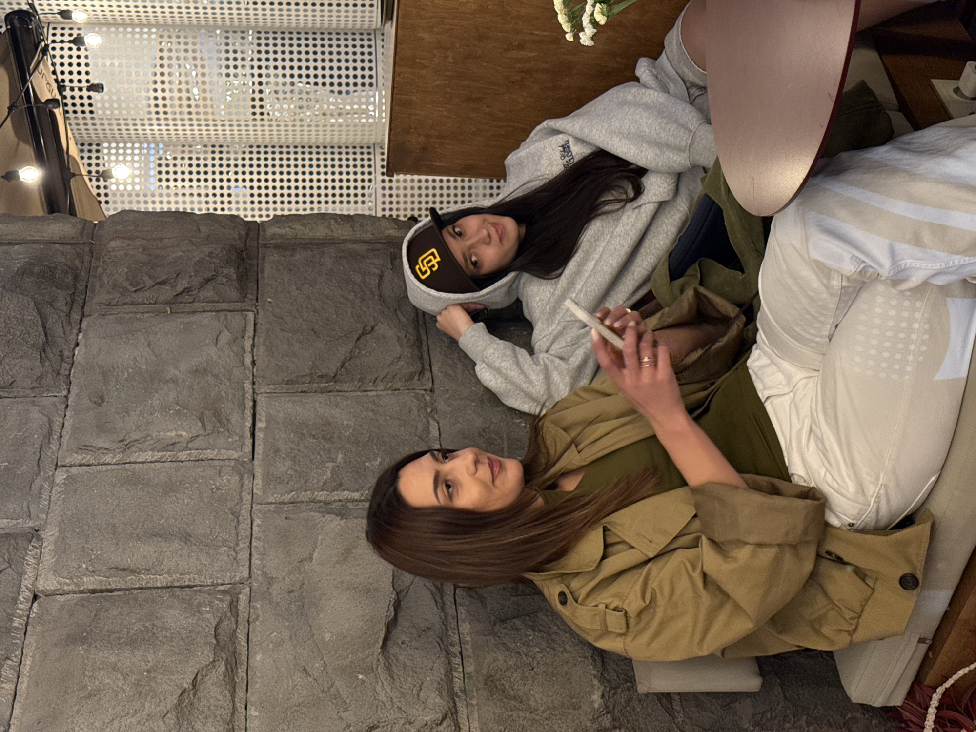 Two women are seated together. One is wearing a grey hoodie and a brown baseball cap. The other is wearing a green shirt and a tan trench coat. Both are holding a phone. They appear to be indoors. The flooring is stone. The ceiling is white and appears to have lights.
