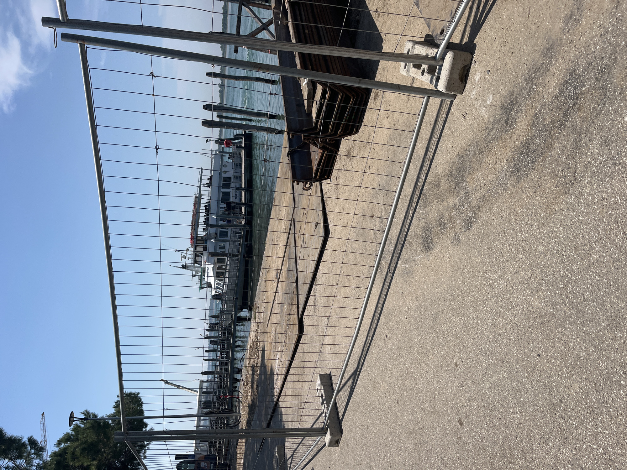 The image shows a fenced-off area at a waterfront location, likely in Punta Sabbioni, Italy. The fence, constructed of metal posts and wire mesh, occupies the foreground and partially obstructs the view. Behind the fence, there is a concrete pier or promenade. Beyond that, the image shows a waterway with boats docked, and a skyline in the distance. A collection of objects that are either rusted or darkly colored are also present. The sky is clear and blue. There are no people visible in the area immediately behind the fence.