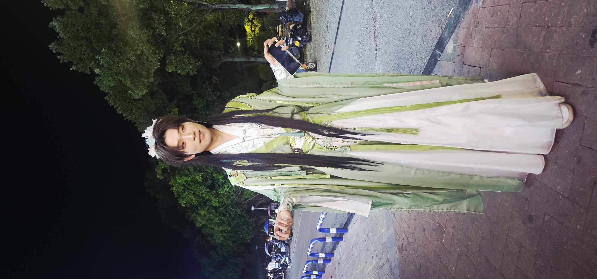 A young East Asian person stands outdoors at night, dressed in a green and white traditional Chinese-style robe with long black hair. They wear a floral headband and hold a smartphone in their right hand. The background features trees, streetlights, parked motorcycles, and a sidewalk with blue and white bollards. The ground is paved with bricks and asphalt. The scene appears to be in the Chongqing High-tech Zone, China, possibly on a street or pathway near an area with parked vehicles.