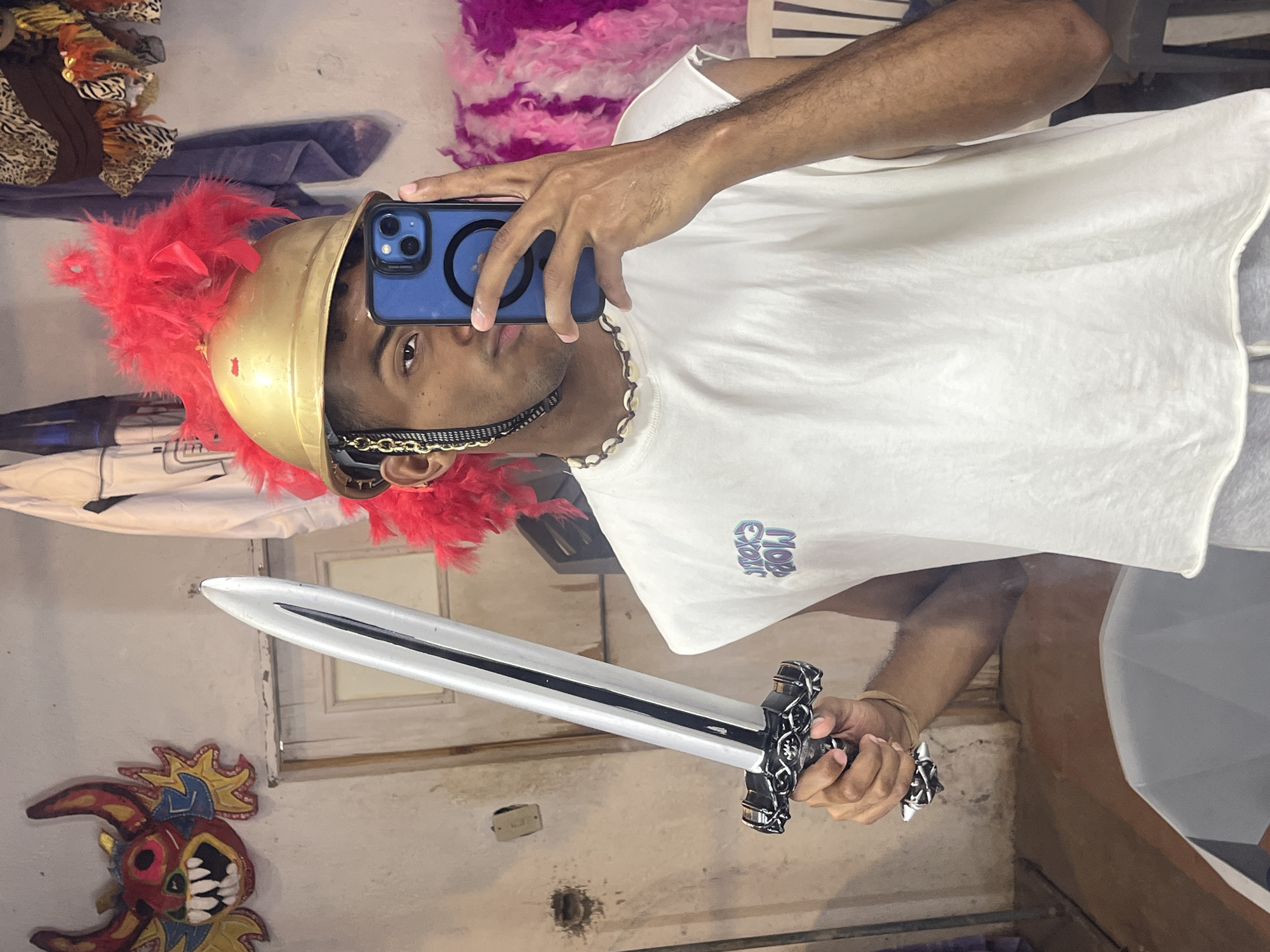 A person wearing a golden Roman helmet with red feathers is taking a selfie in front of a mirror, holding a toy sword. The person is wearing a white t-shirt and a shell necklace. A blue iPhone is used to take the picture. The background shows a cluttered room with clothes, a pink feather boa, and a decorative mask hanging on the wall. The location is possibly a private residence, but there is no information to confirm the exact city or country, specifically Porlamar, Venezuela.