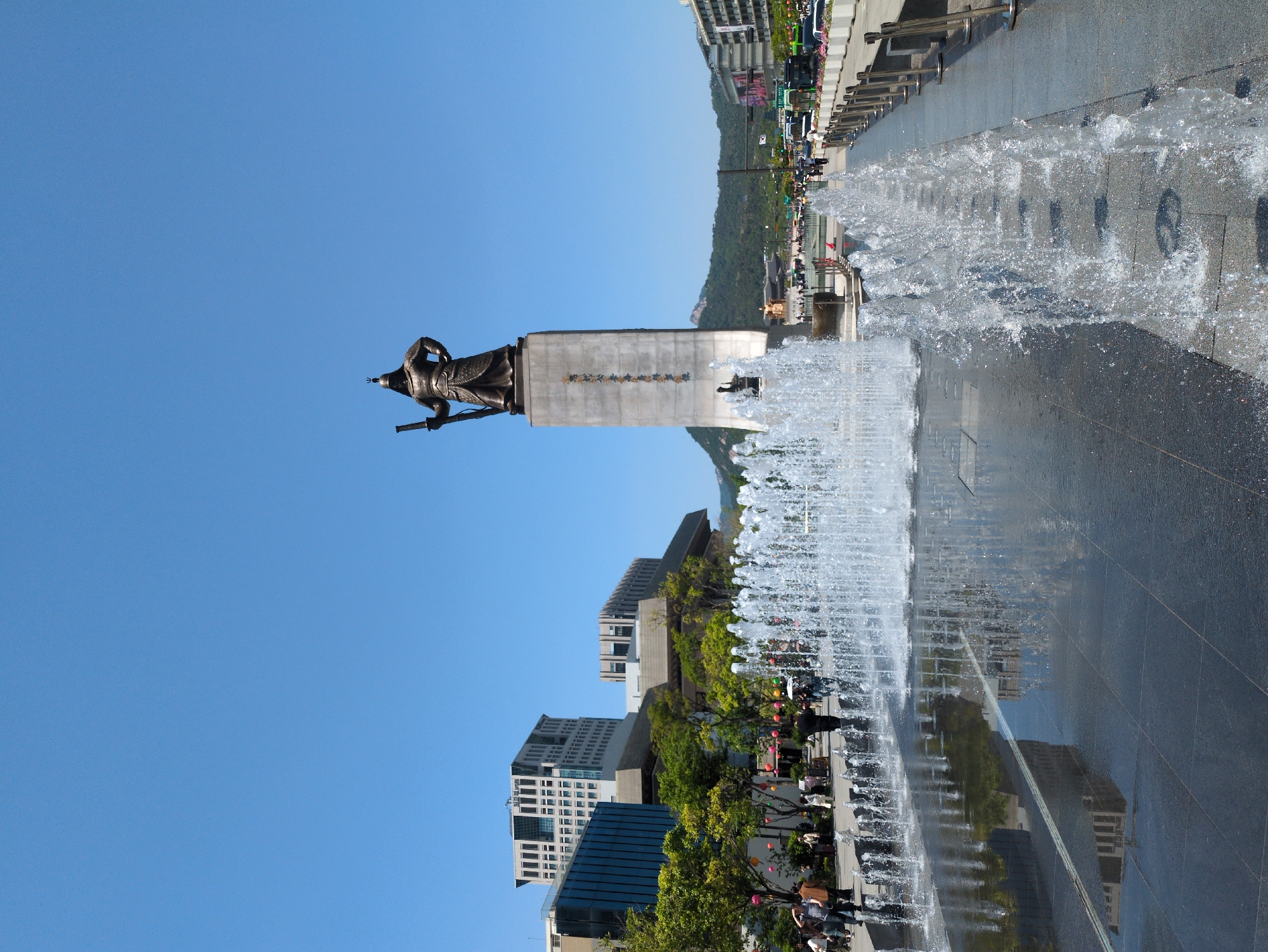 The image shows the Statue of Admiral Yi Sun-sin in Seoul, South Korea. The bronze statue stands on a stone plinth in Gwanghwamun Square, a public plaza in the heart of the city. In front of the statue is a water fountain with multiple jets spraying water upwards, creating a visual spectacle. The ground is paved with dark stone tiles, reflecting the surrounding buildings and the sky. Buildings of varying heights and architectural styles are visible in the background, indicating a modern urban environment. Beyond these, there's a glimpse of a tree-covered hill or mountain. The sky is clear and blue, suggesting a sunny day. On the side, water is spraying onto a walkway. People are seen walking along the side of the square, some possibly looking at the statue or the fountain.
