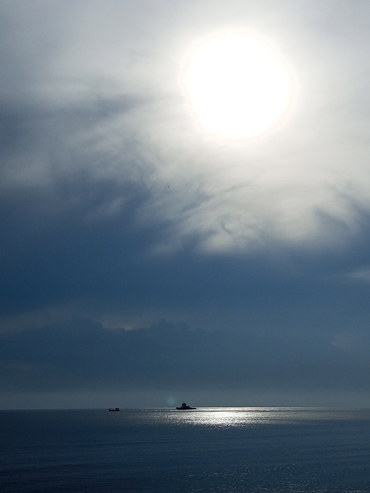 The image shows a seascape under a bright, overcast sky. The sun is high and obscured by clouds, creating a diffused, circular glow. Below the clouds, the sea is a dark blue, reflecting the light from the sun in a shimmering path. Two small vessels are visible in the distance, silhouetted against the horizon. The location is possibly Lhokseumawe, Indonesia.