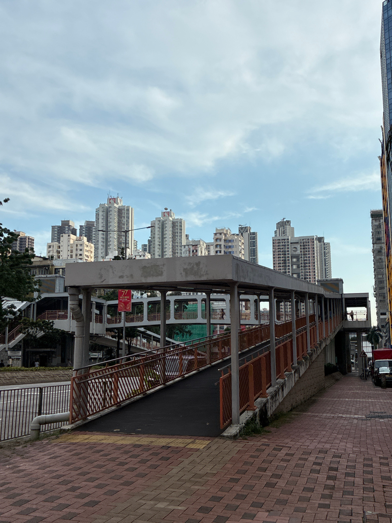 The image depicts a pedestrian overpass in a dense urban environment, likely Hong Kong, China. The overpass, constructed with concrete and featuring orange metal railings, spans across a street with a brick-patterned sidewalk in the foreground. In the background, a cluster of high-rise apartment buildings, characteristic of Hong Kong's cityscape, are visible under a partly cloudy sky. The overpass appears to be a functional piece of city infrastructure, with a dark asphalt walkway leading up to it. A few individuals are discernible on the overpass and in the background, suggesting everyday urban activity. The overall scene captures the verticality and density of a Hong Kong neighborhood.