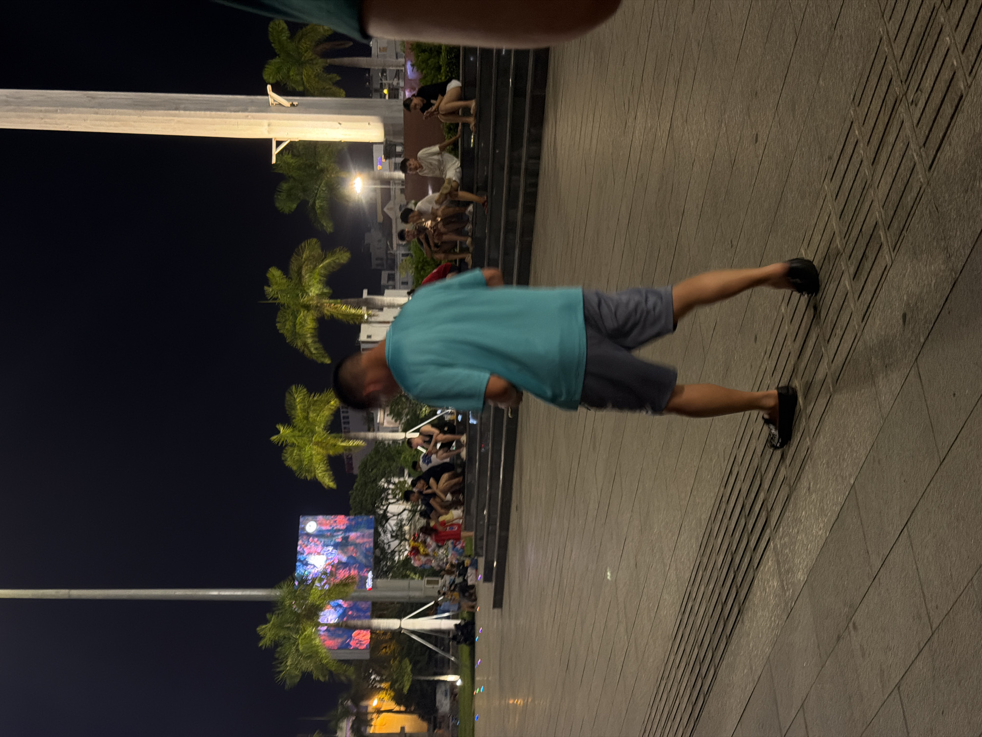 This is a nighttime photograph taken at the Dragon Bridge plaza in Da Nang, Vietnam. In the foreground, a man in a teal t-shirt and gray shorts is walking away from the camera, holding a phone. He is wearing black slip-on shoes. The ground is paved with large rectangular tiles and has a textured strip running parallel to the man's path. In the midground, a group of people are seated on wide concrete steps. They are casually dressed and appear to be looking at their phones or talking amongst themselves. Behind the steps, several palm trees are illuminated, and a large digital billboard displays a colorful, abstract image. In the background, a tall, white pillar, possibly part of a structure or monument, is visible. The overall atmosphere suggests a public gathering space at night, with artificial lighting from the billboard, streetlights, and possibly decorative ground lighting creating a festive or relaxed ambiance.